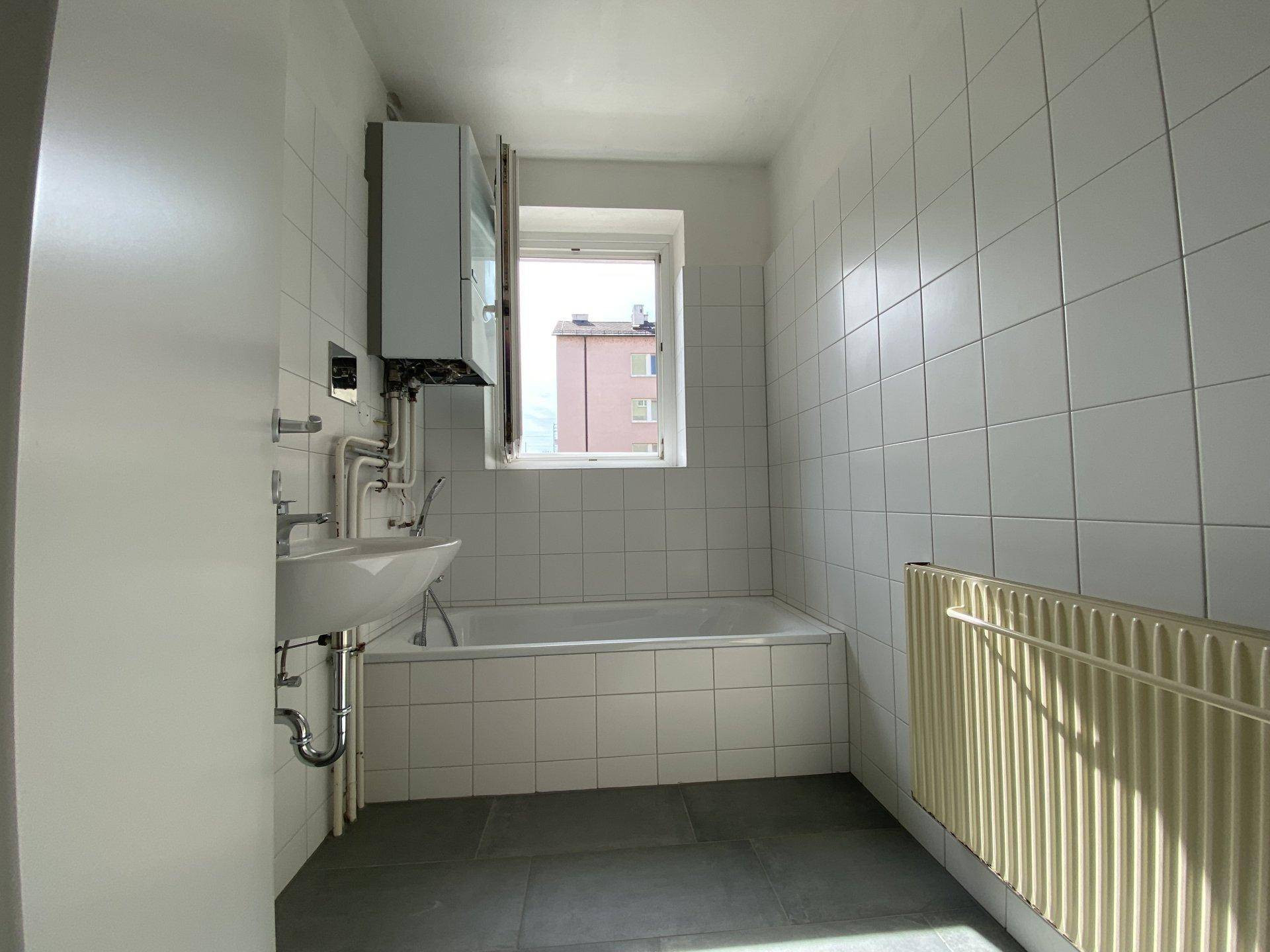 Badezimmer mit weißen Fliesen, Badewanne, Waschbecken und Fenster.