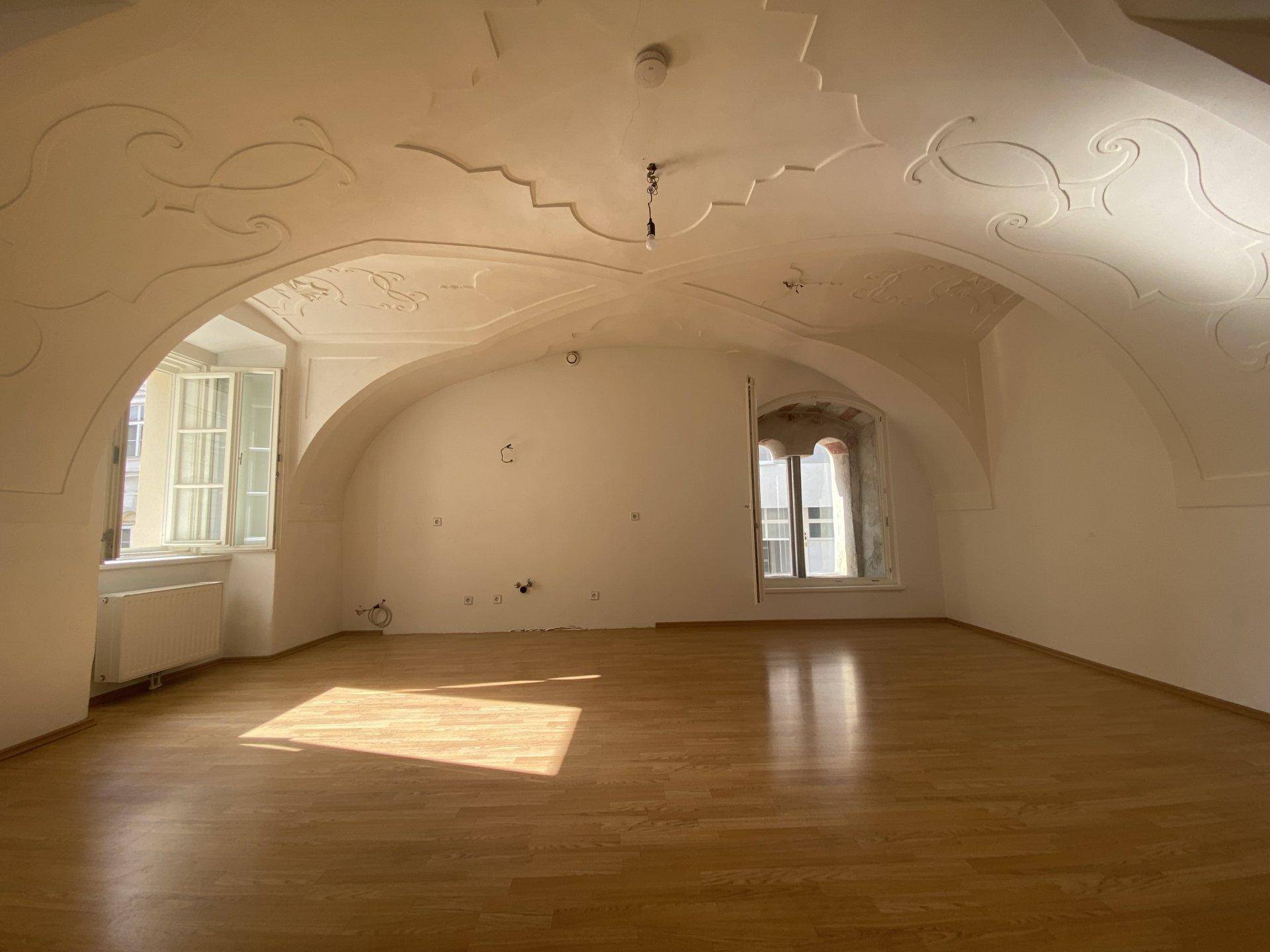 Geräumiges Wohnzimmer mit beeindruckender Stuckdecke, gewölbten Fenstern und Holzboden.