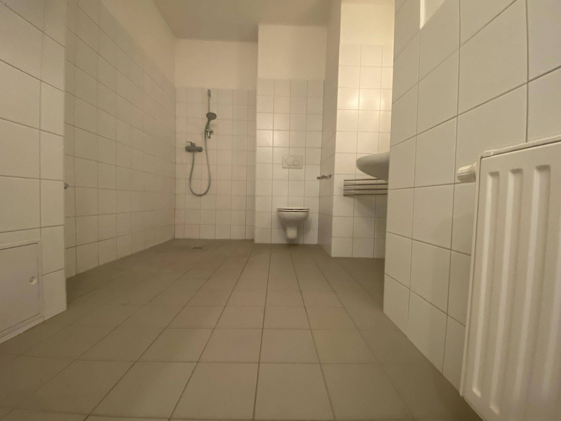 Einfaches Badezimmer mit weißen Fliesen, Dusche und wandhängendem WC.