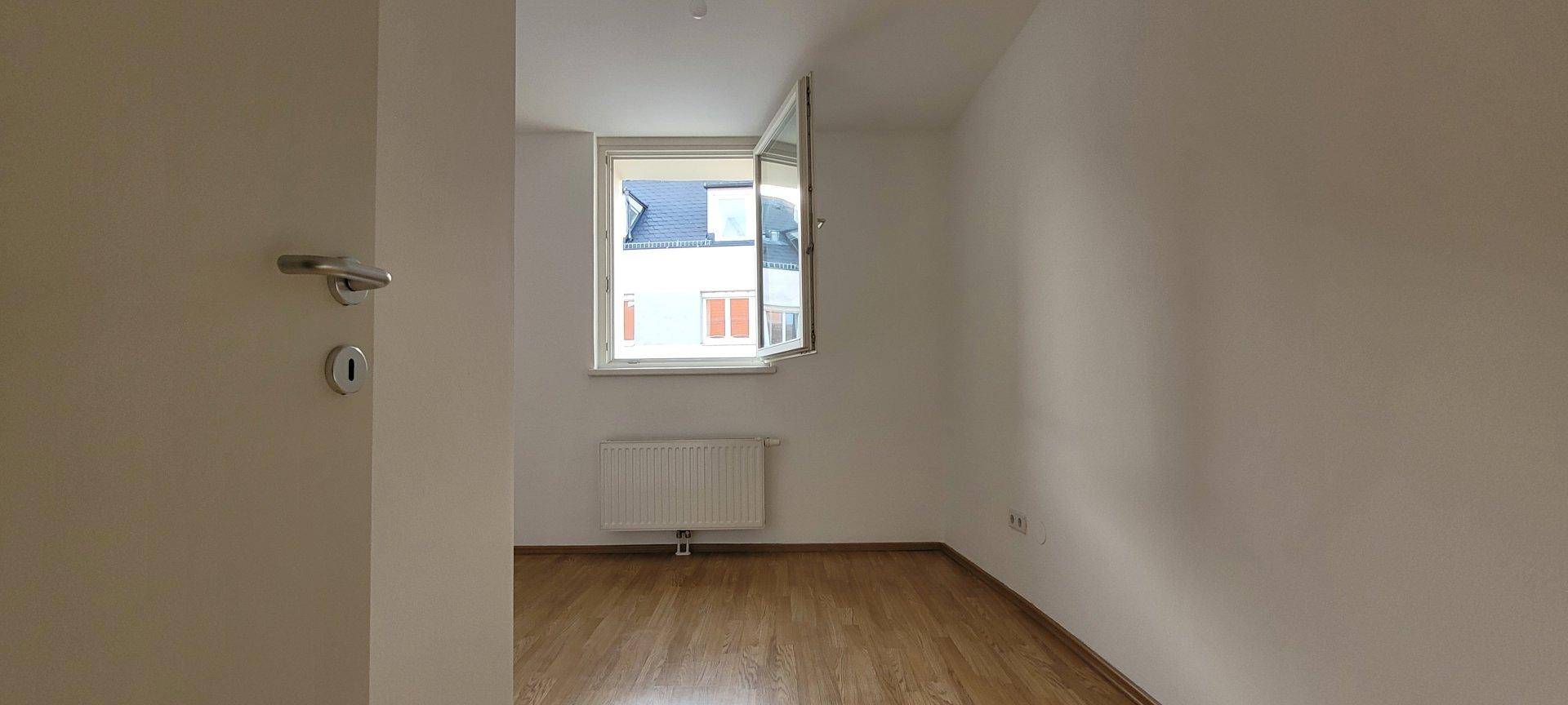 Leeres Schlafzimmer mit Holzboden und einem Fenster, das viel Licht hereinlässt.