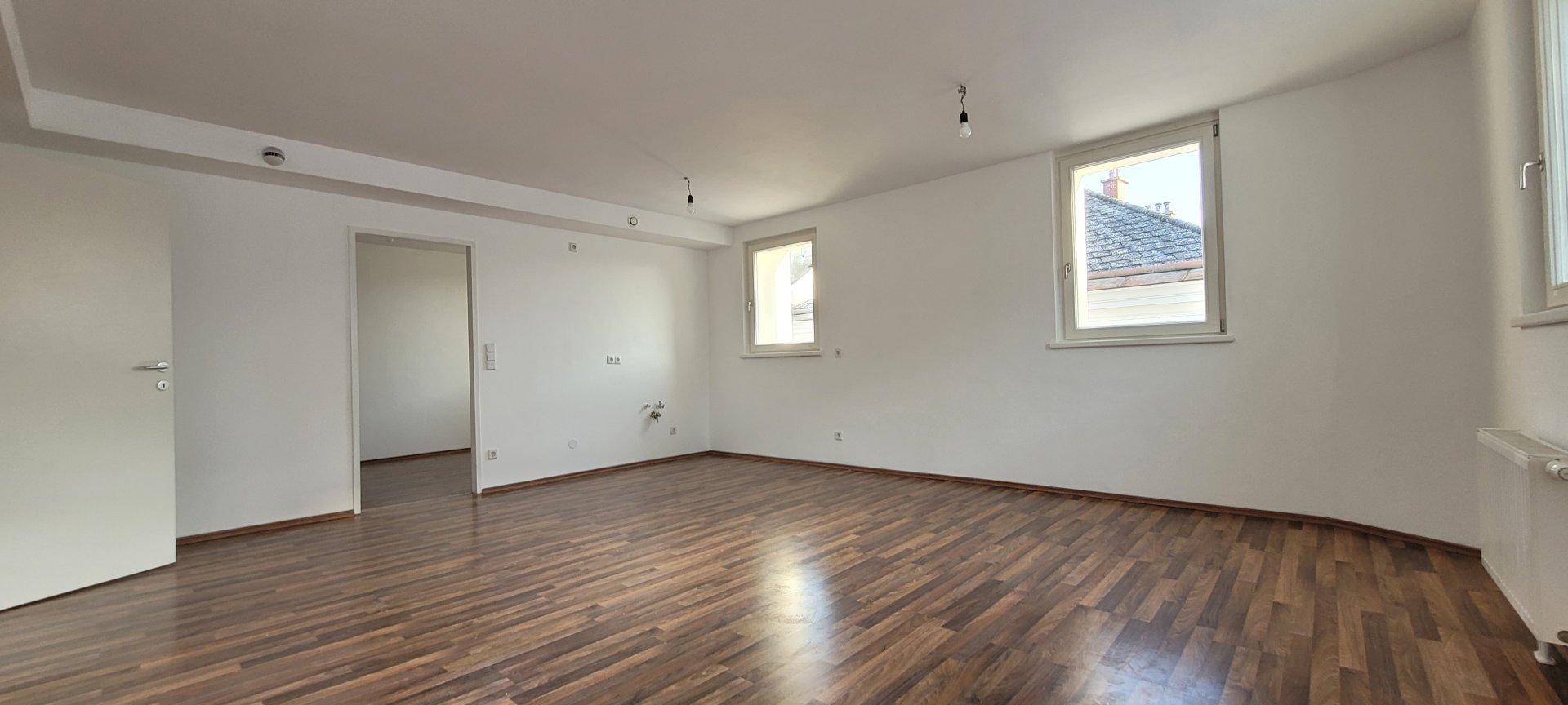 Geräumiges, leeres Wohnzimmer mit Holzboden, weißen Wänden und zwei Fenstern.