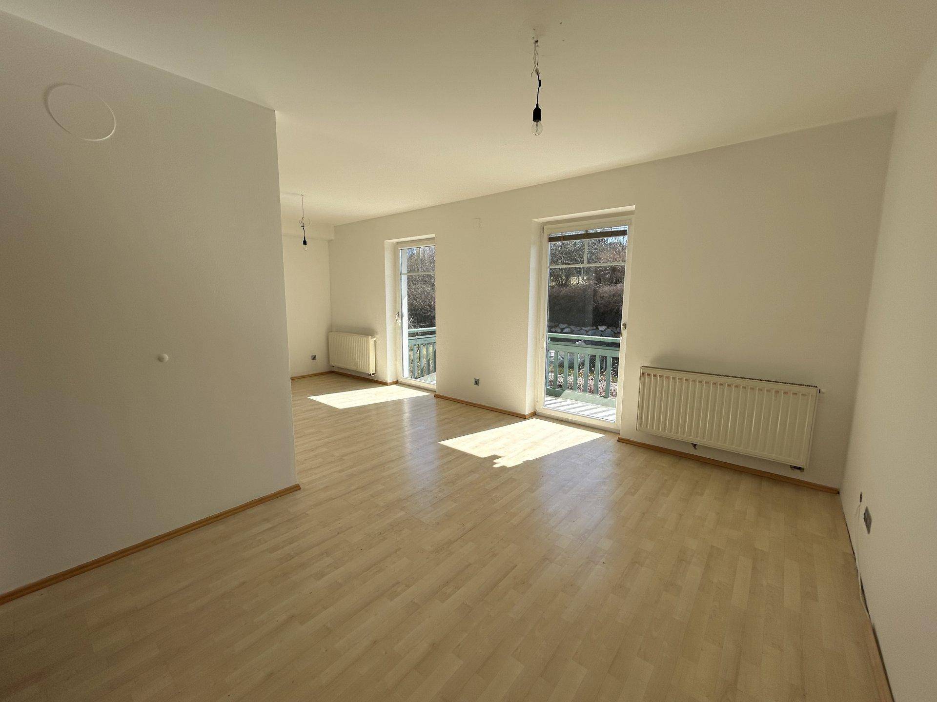 Geräumiges Wohnzimmer mit Laminatboden, großen Fenstern und Zugang zum Balkon.