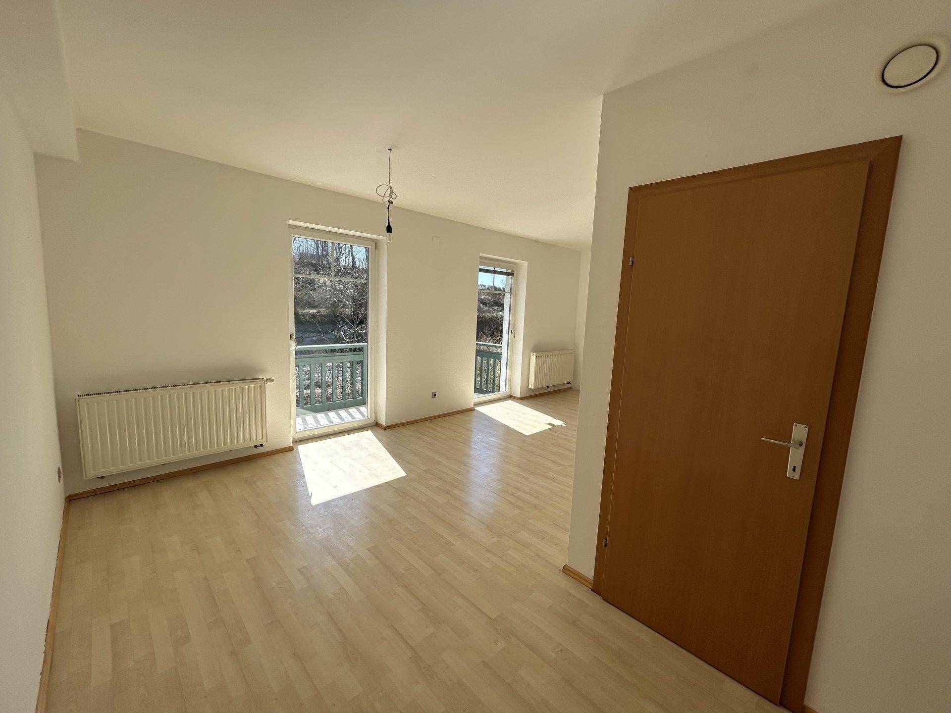 Sonniger Wohnraum mit Laminatboden und zwei Fenstern, die viel Tageslicht hereinlassen.
