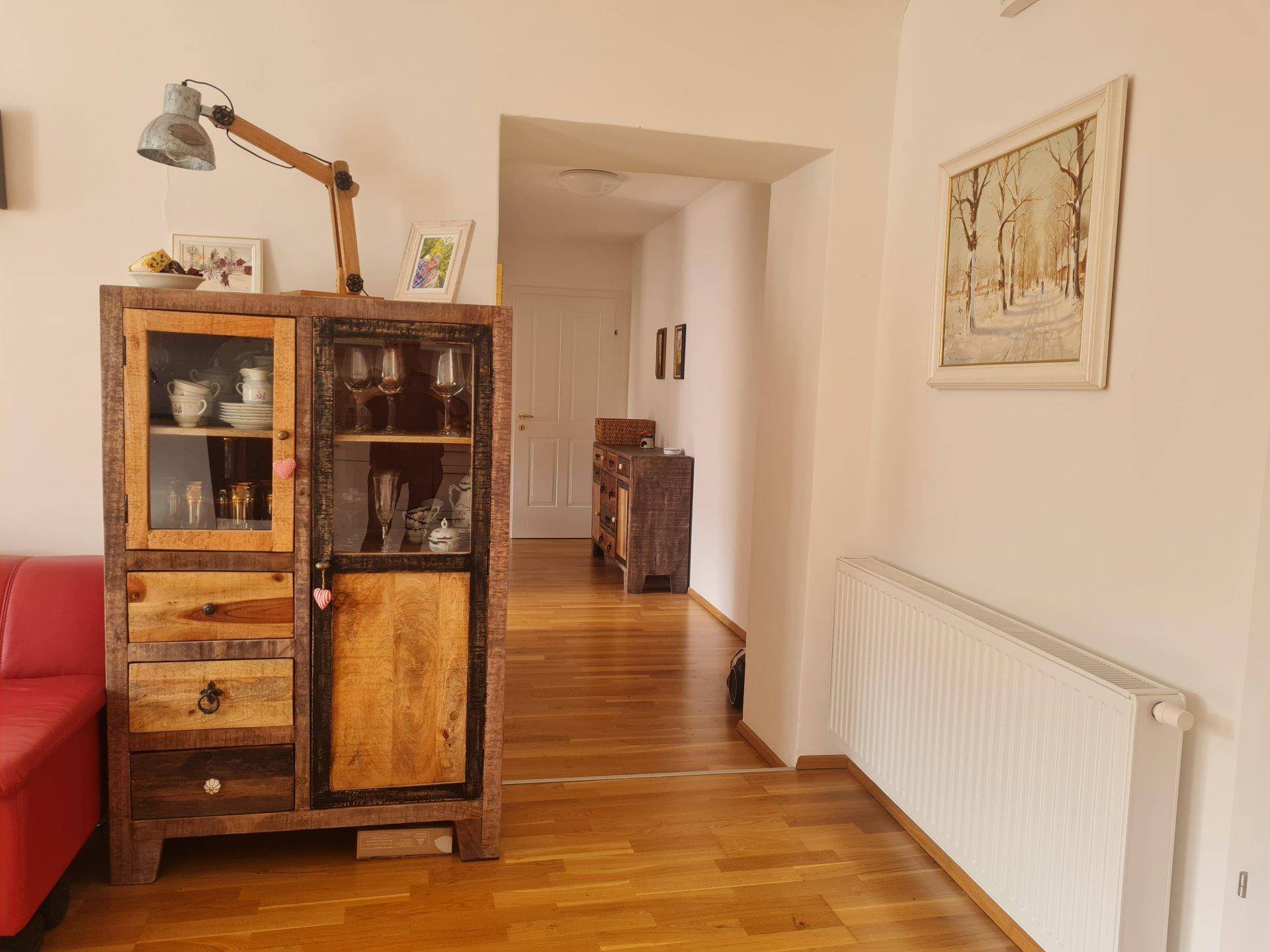 Blick vom Wohnbereich in den Flur mit einem rustikalen Holzschrank und Heizkörper.
