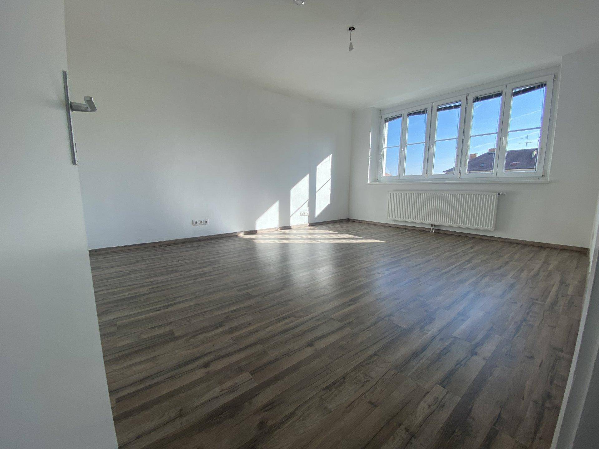Geräumiger, heller Raum mit Laminatboden, weißen Wänden und einem großen Fenster mit Heizkörper.