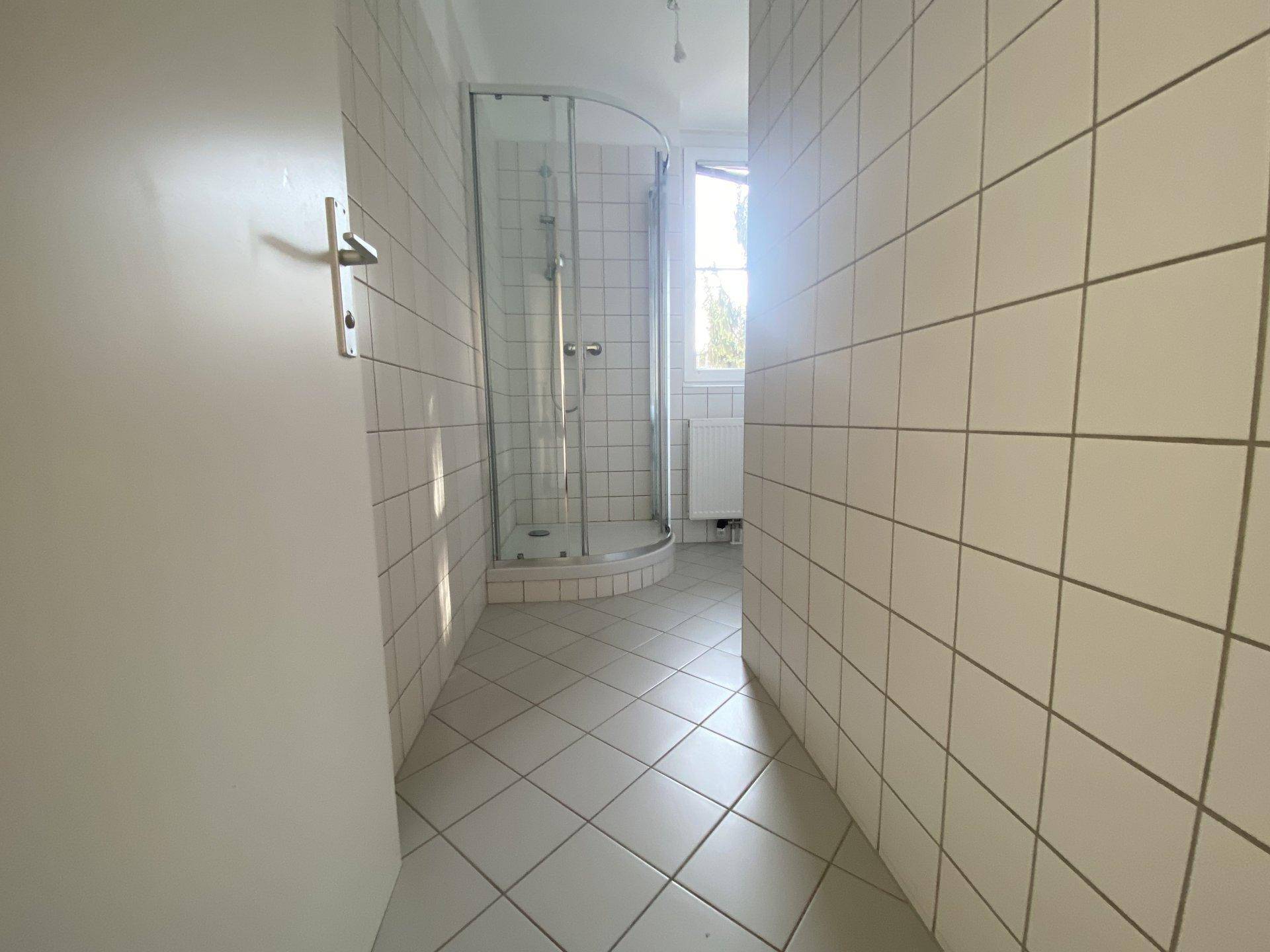Helles Badezimmer mit weißen Fliesen an Wänden und Boden sowie einer runden Duschkabine und Fenster.