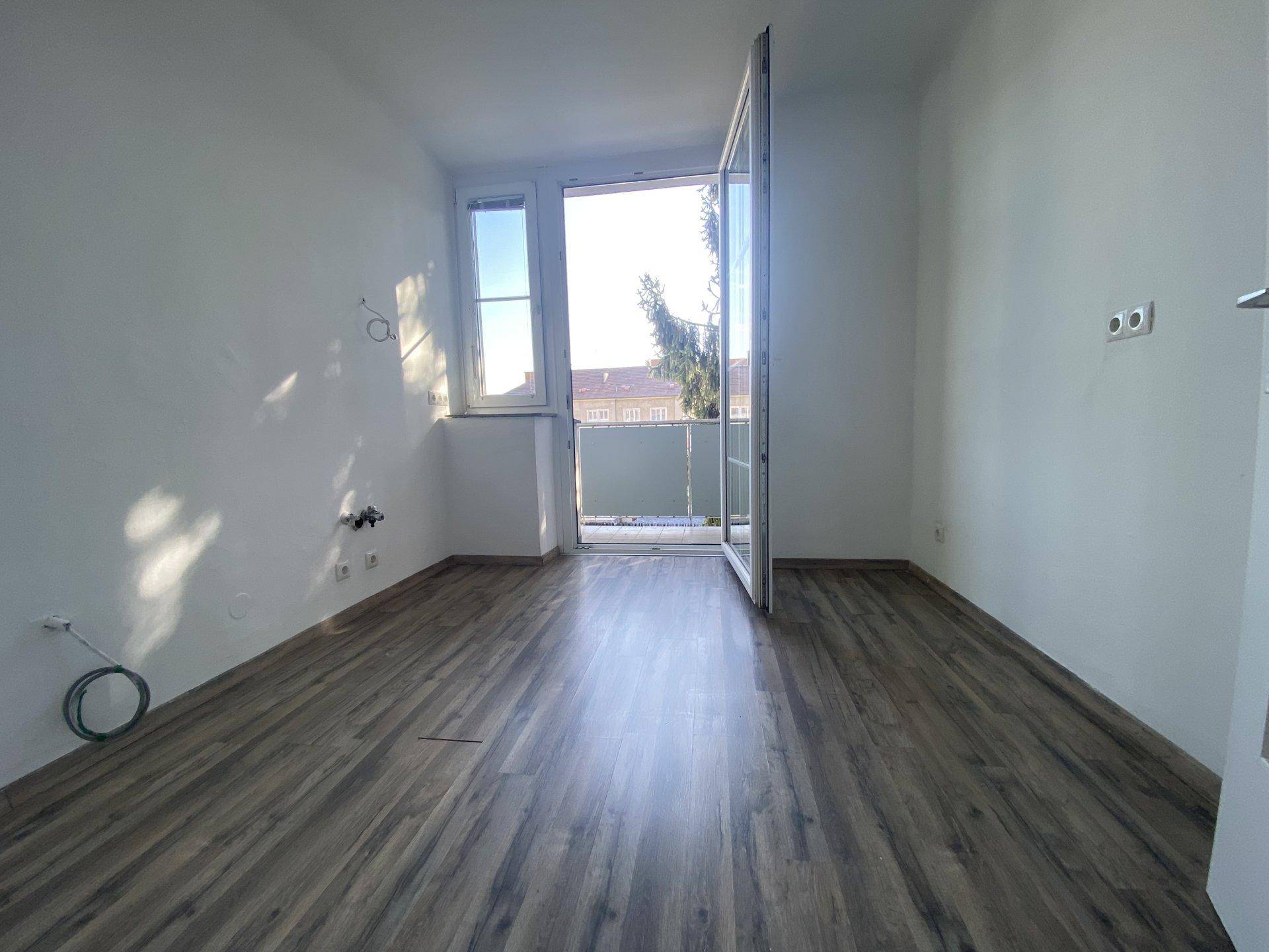 Leerer Raum mit Laminatboden, weißen Wänden und Zugang zu einem Balkon, der viel Tageslicht bietet.