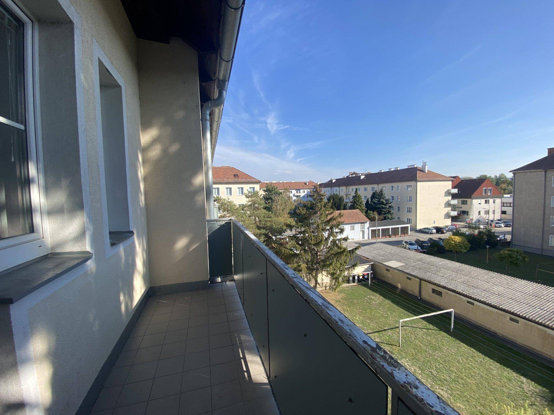 Balkon mit Blick auf eine Wohngegend, Bäume und einen kleinen Rasenplatz unter blauem Himmel.