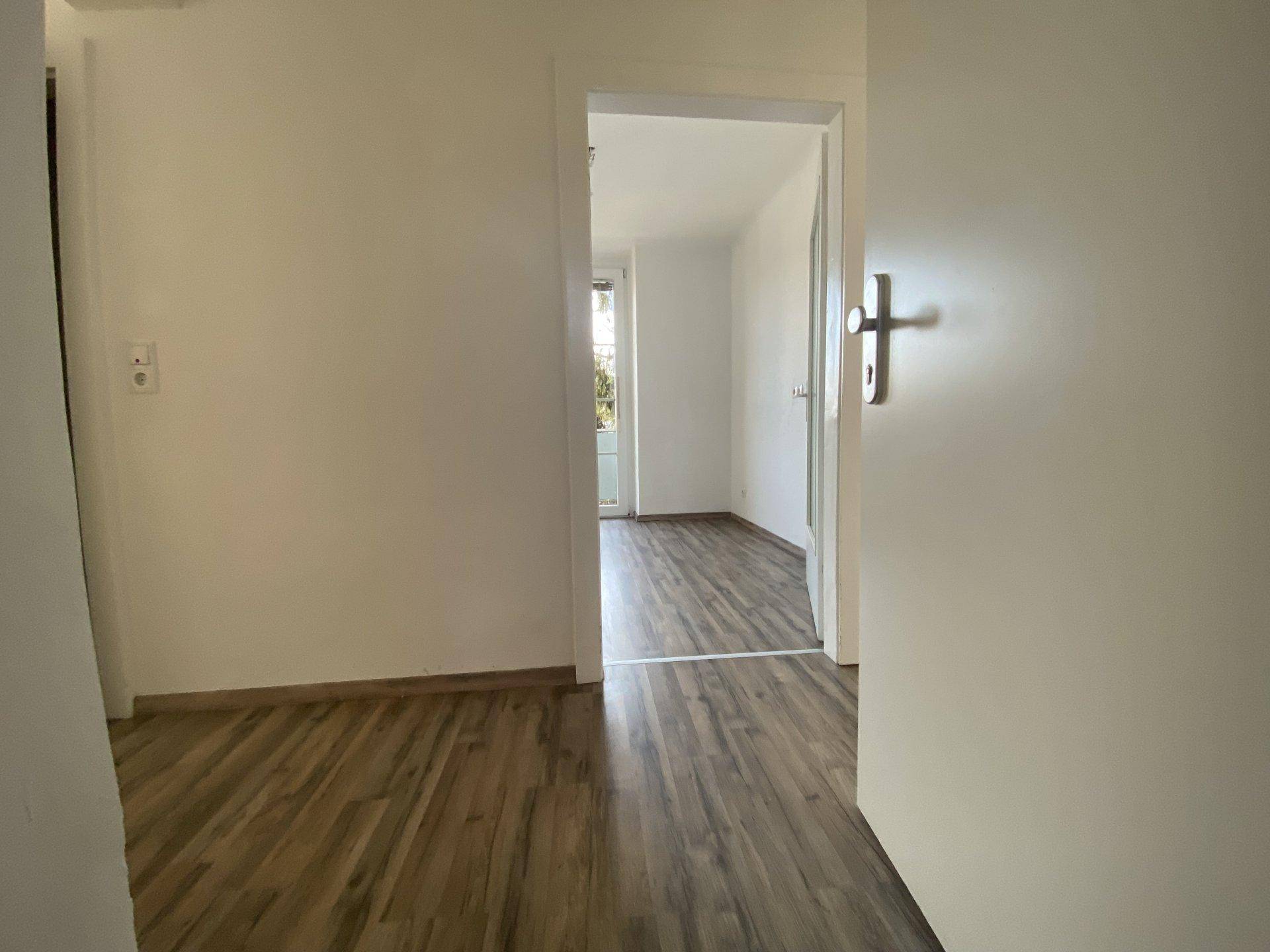 Heller Flur mit Laminatboden und weiß gestrichenen Wänden, der in einen weiteren Raum führt.