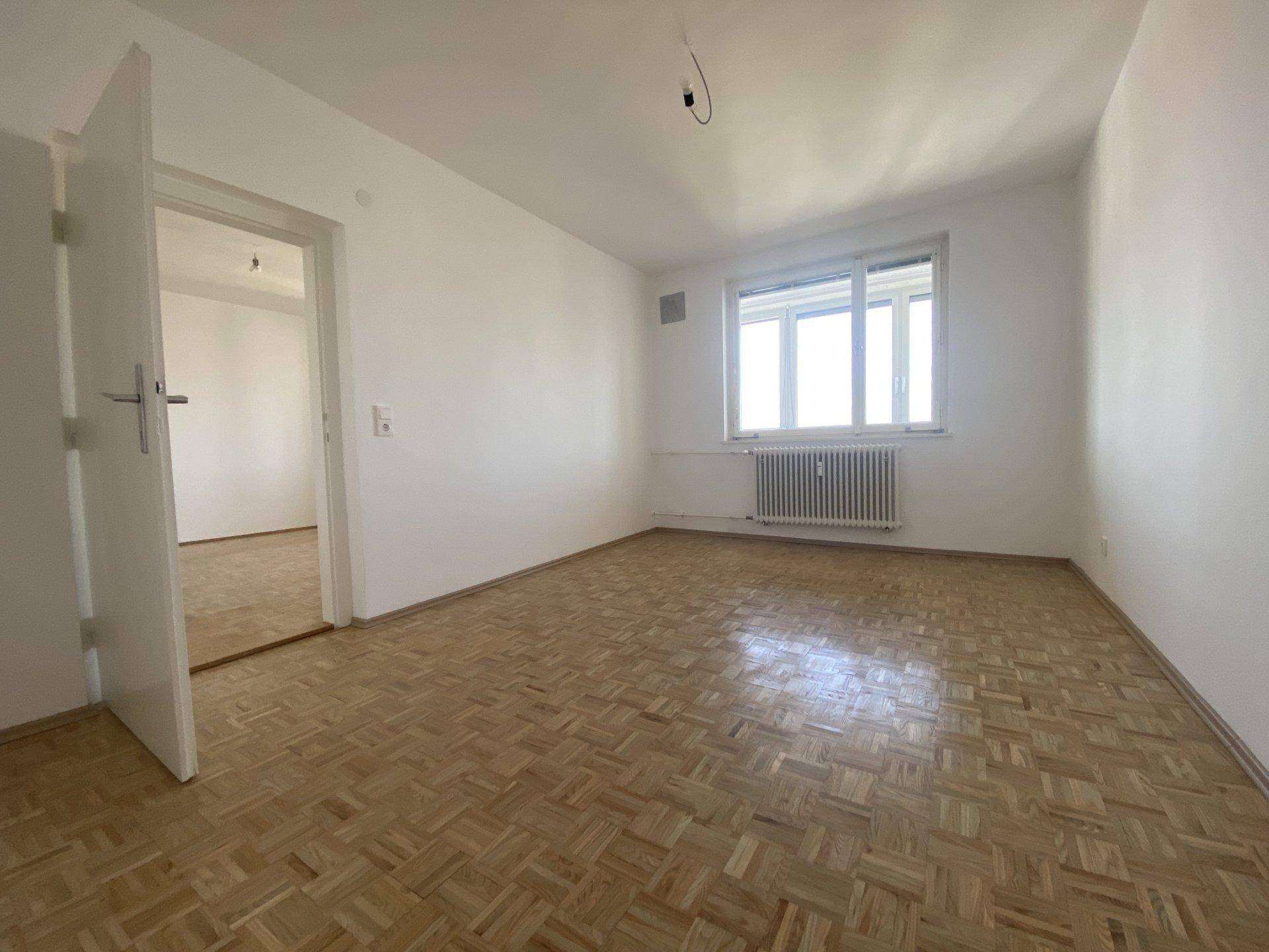 Leeres Zimmer mit Parkettboden, großem Fenster und Heizkörper, bereit zur individuellen Gestaltung.