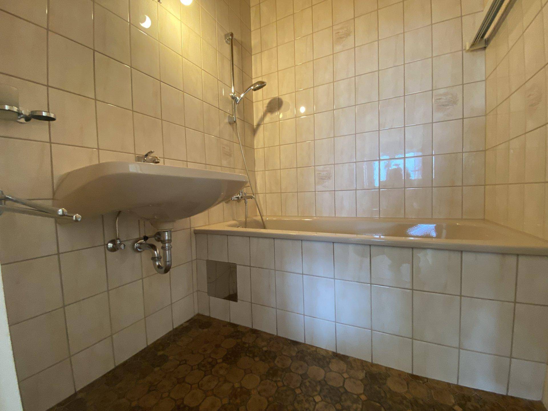 Badezimmer mit Badewanne, Waschbecken und hellen Fliesen, ausgestattet mit einer Duschfunktion.