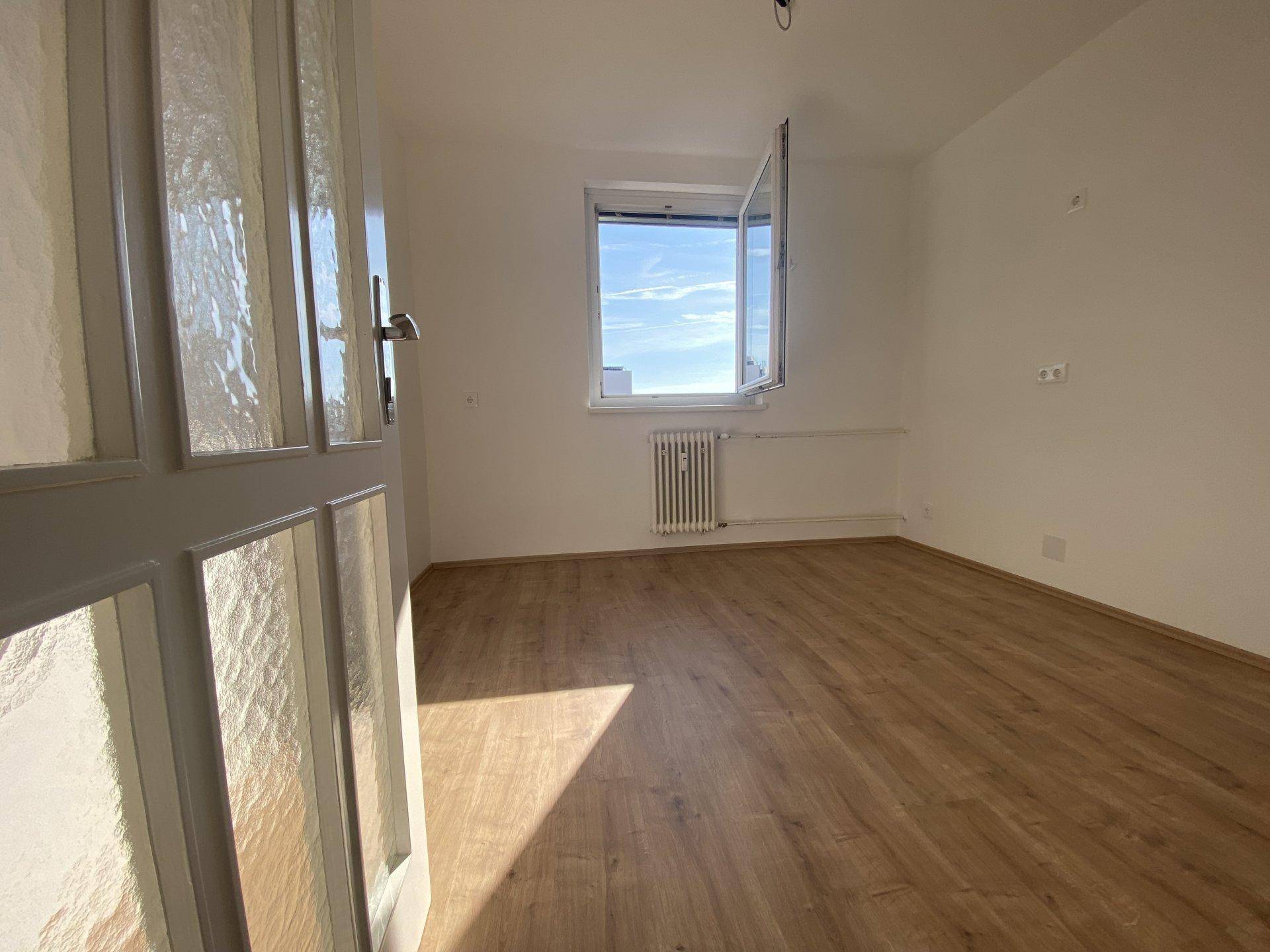 Heller, leerer Raum mit Laminatboden und einem Fenster, das viel Tageslicht hereinlässt.