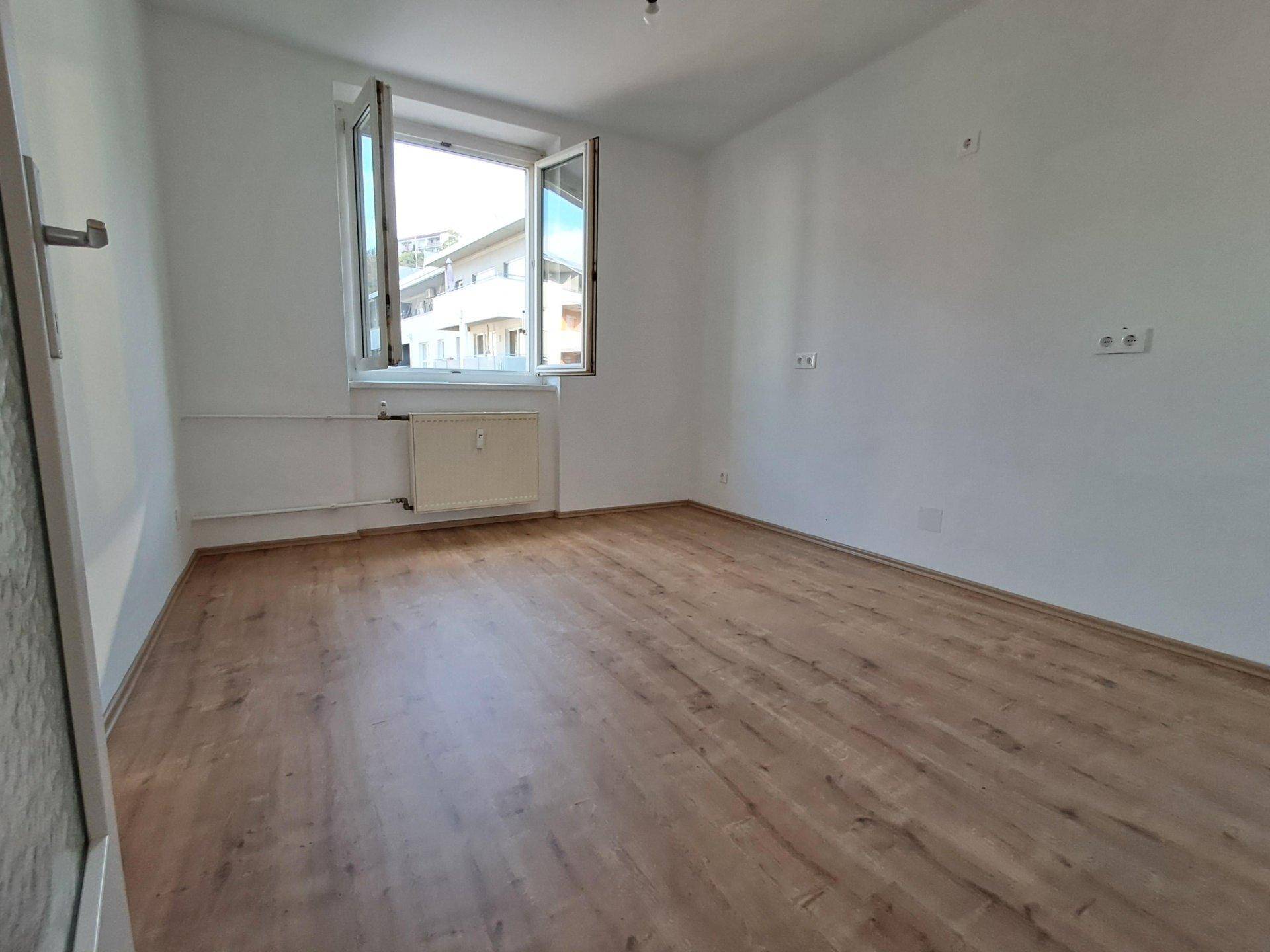 Leeres Zimmer mit Laminatboden, weiß gestrichenen Wänden und einem Fenster.