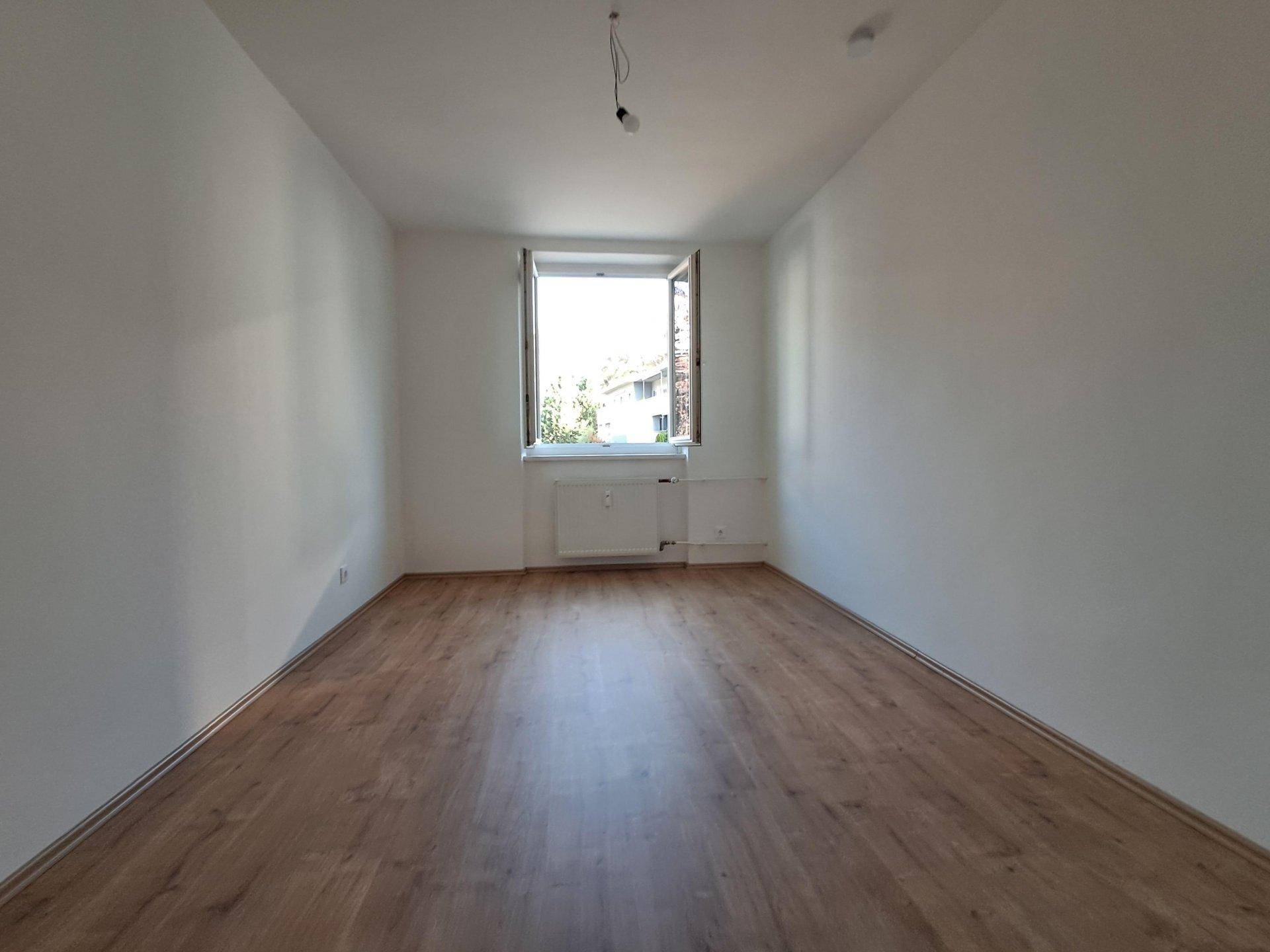 Helles, leeres Zimmer mit Laminatboden und einem Fenster, das viel Licht hereinlässt.