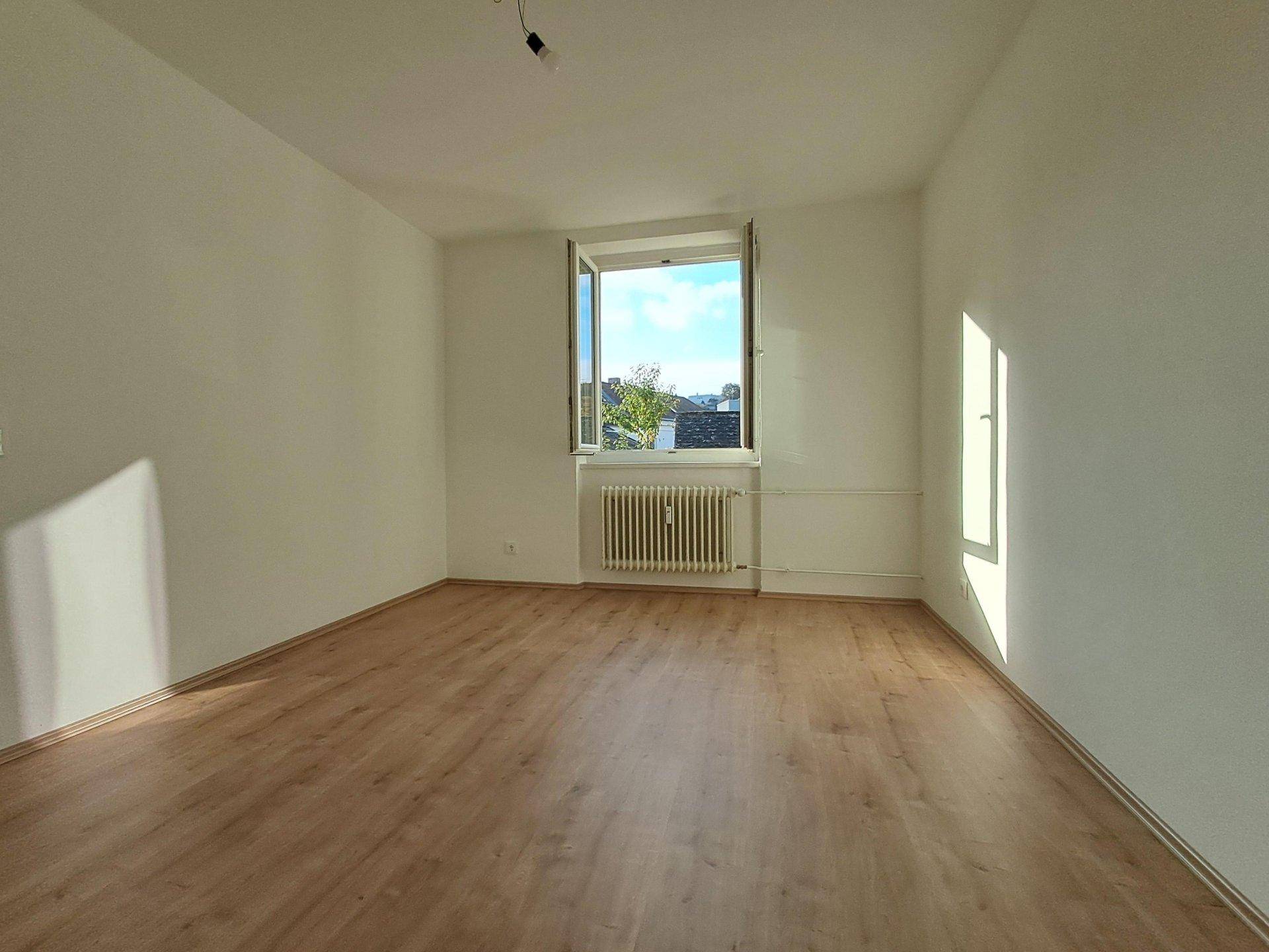 Freundliches Zimmer mit Laminatboden, weiß gestrichenen Wänden und einem großen Fenster.