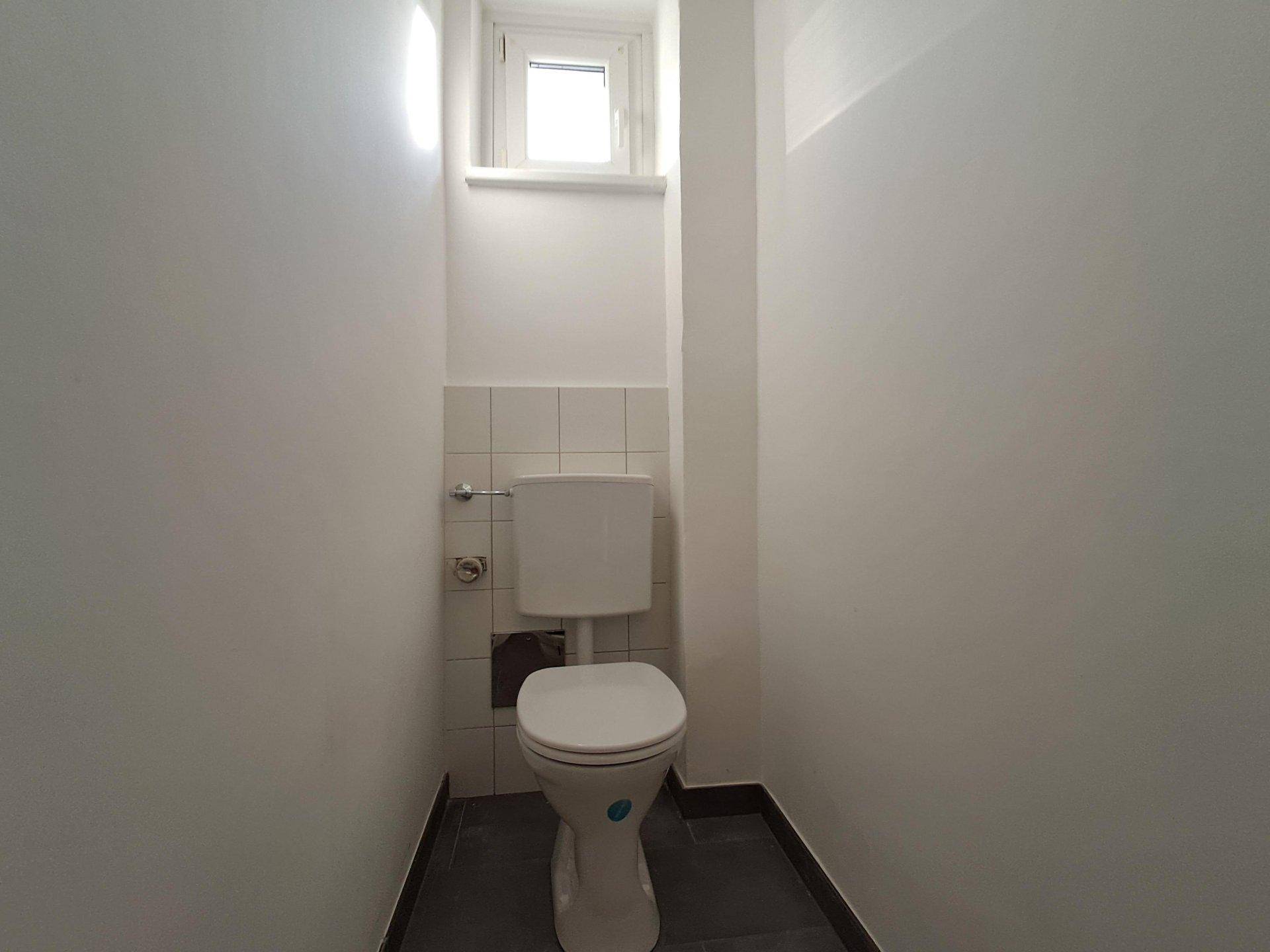 Separate Toilette mit Fenster, weißer Keramik und dunklem Fliesenboden.