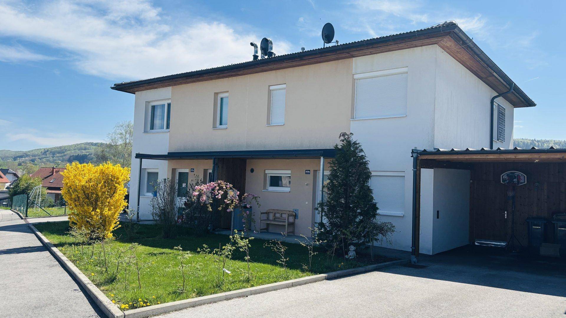 Ansicht der Doppelhaushälfte mit blühendem Strauch und Carport an einem sonnigen Tag.