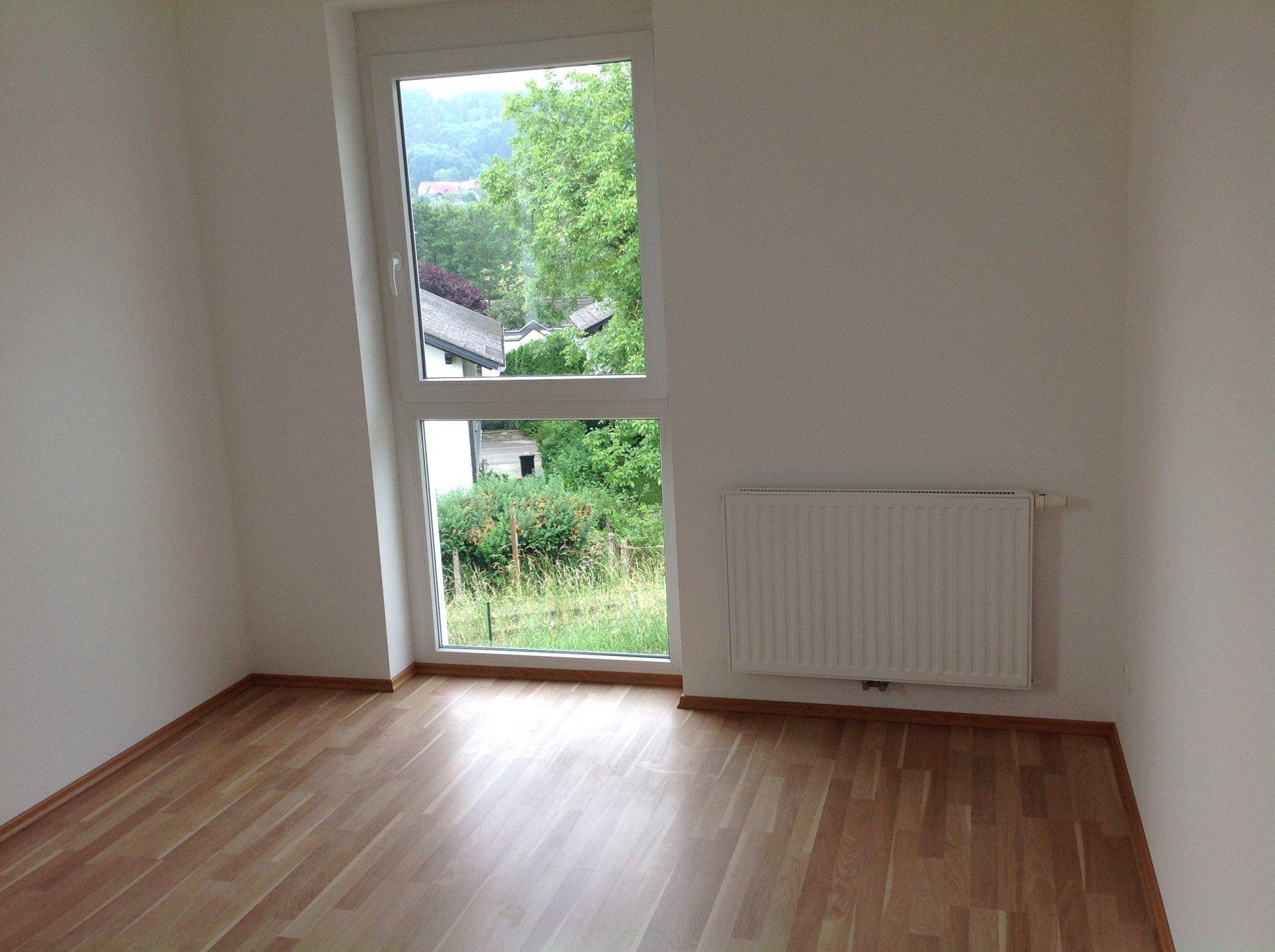 Helles Zimmer mit Laminatboden, Heizkörper und hohem Fenster mit grüner Aussicht.