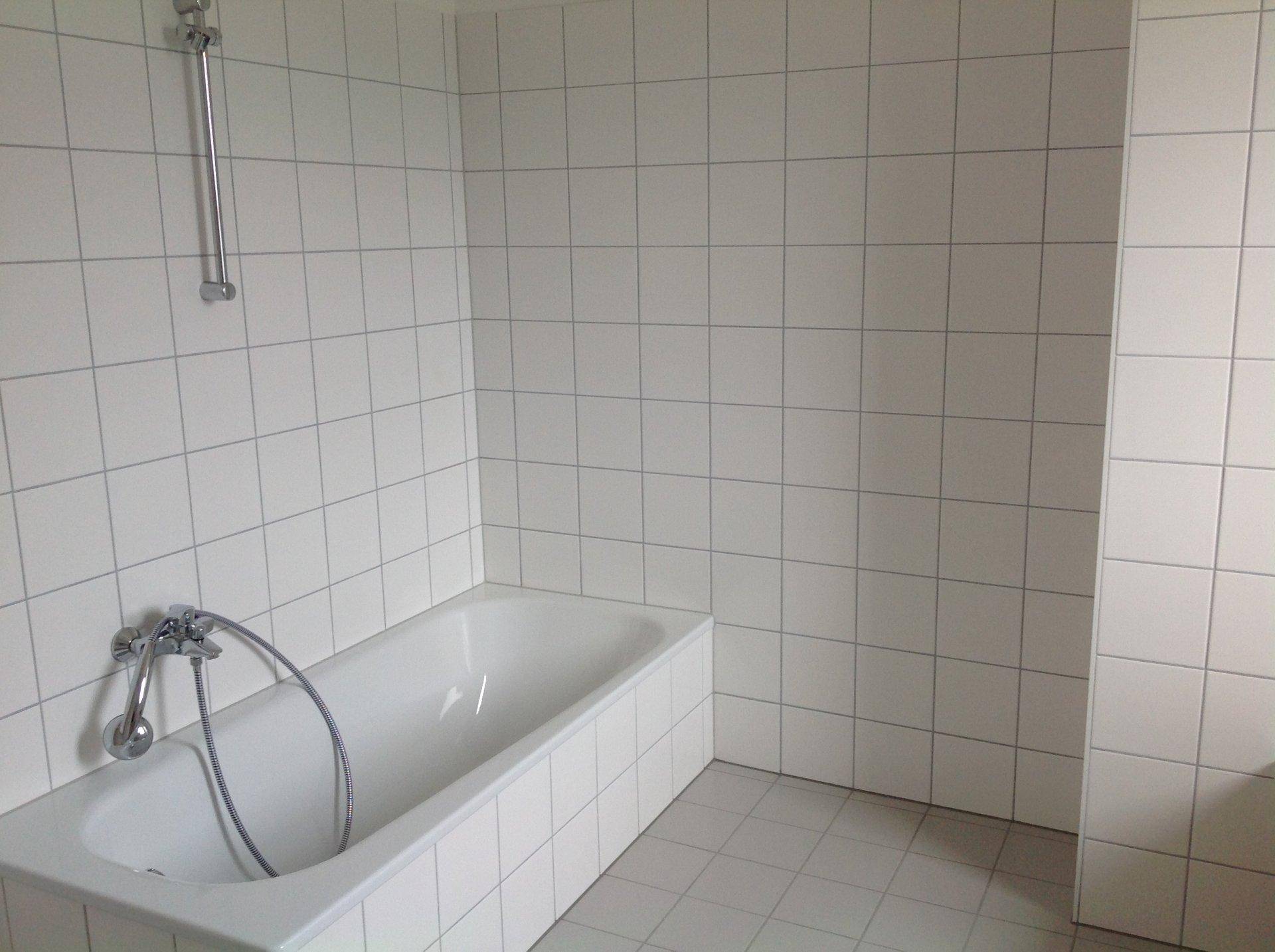 Badezimmer mit weißen Wand- und Bodenfliesen sowie einer Badewanne mit Duscharmatur.