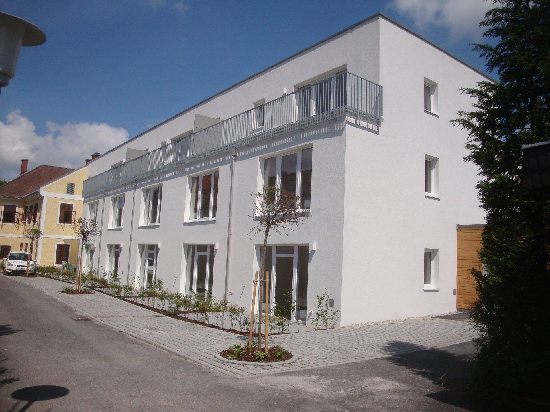 Modernes weißes Gebäude mit klaren Linien, großen Fenstern und Balkonen an einem sonnigen Tag.