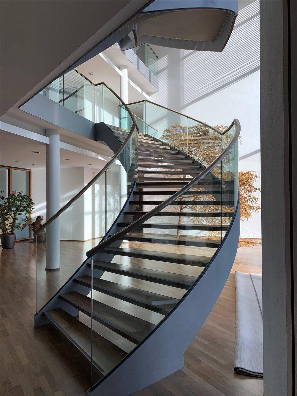 Elegante geschwungene Treppe mit Glasgeländer und Holzstufen, die viel natürliches Licht empfängt.