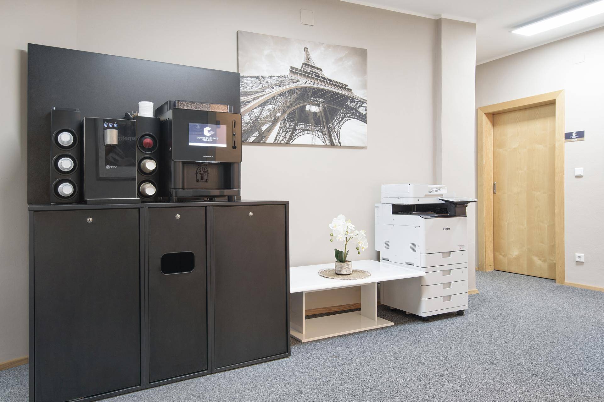 Kleine Lounge-Ecke mit Kaffeemaschine, Drucker und einem Bild des Eiffelturms an der Wand.