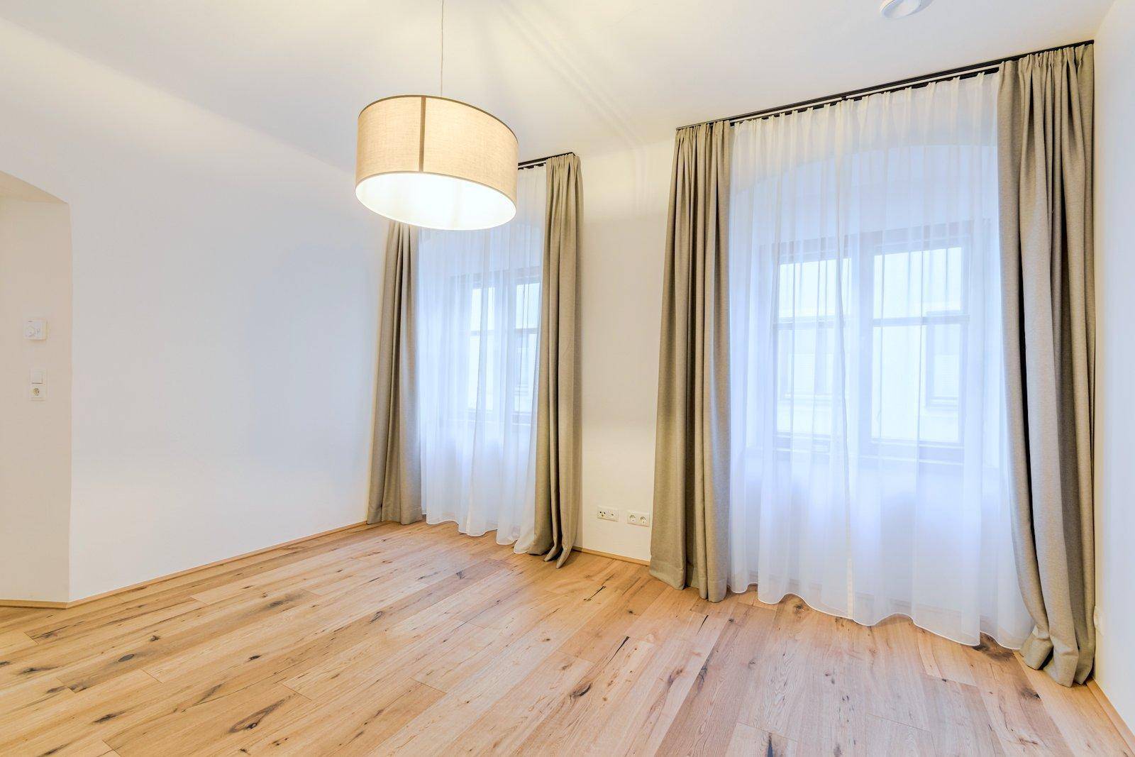 Helles, leeres Zimmer mit Holzboden, weißen Wänden und zwei Fenstern mit Vorhängen sowie einer modernen Deckenleuchte.