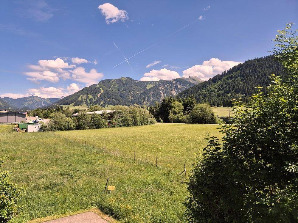 Weitläufiger Blick über grüne Wiesen und bewaldete Berge unter blauem Himmel.