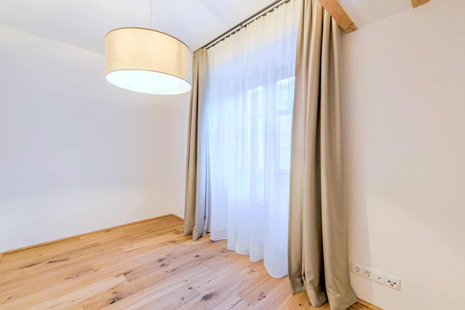 Helles, leeres Zimmer mit Holzboden, weißen Wänden, einem Fenster mit Vorhängen und einer modernen Deckenlampe.