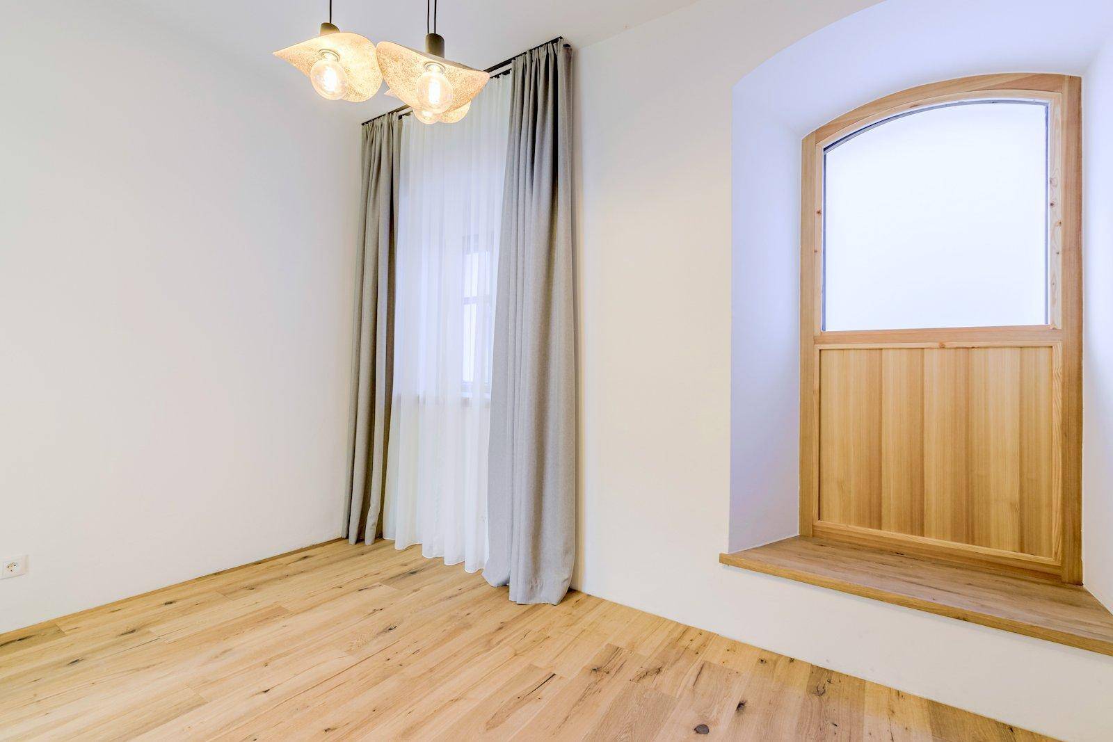 Heller Raum mit Holzboden, Vorhängen und einem besonderen Holzbogenfenster, das viel Licht hereinlässt.