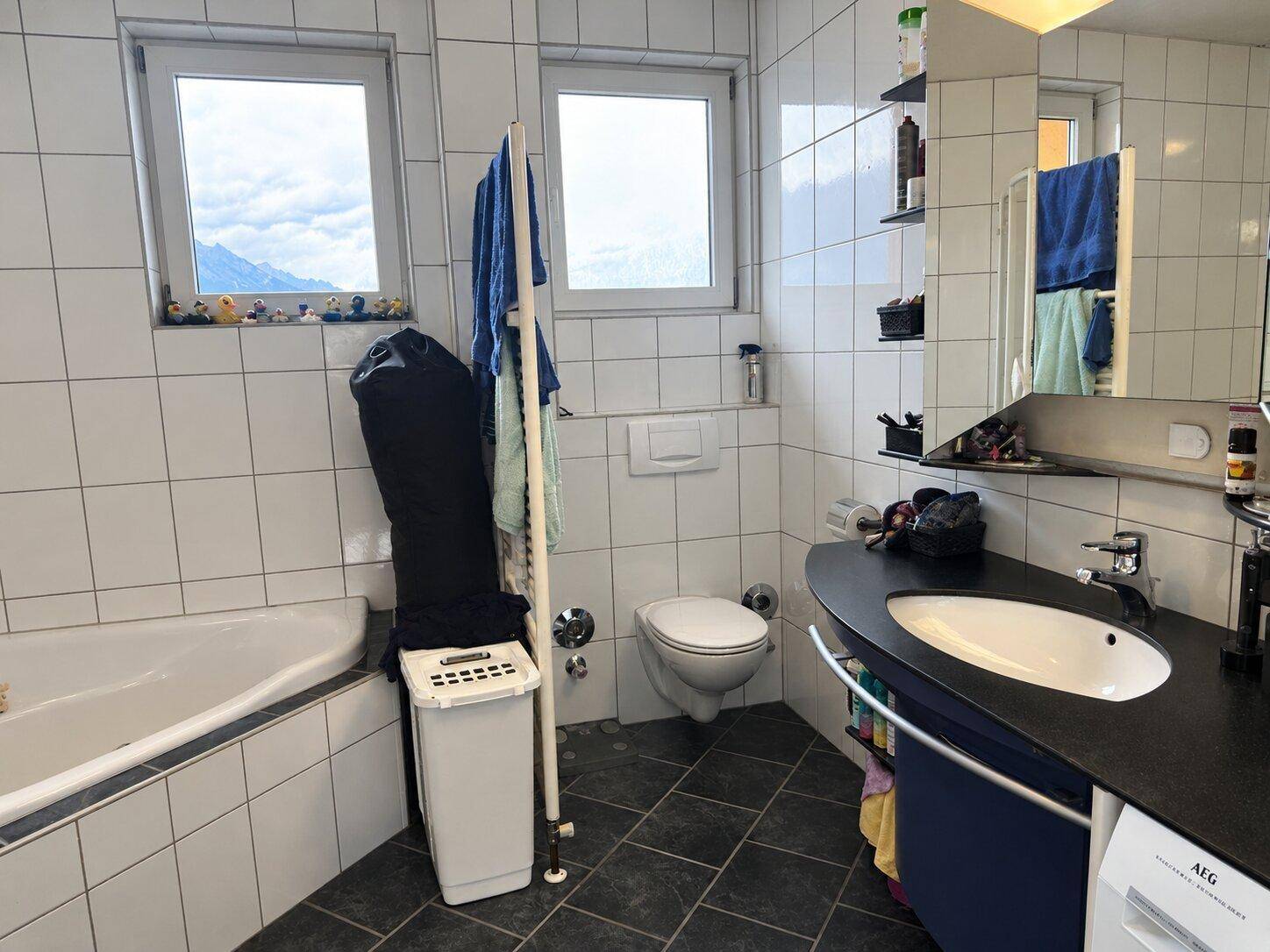 Badezimmer mit weißen Fliesen, WC, Waschtisch und Fenstern mit Bergblick.