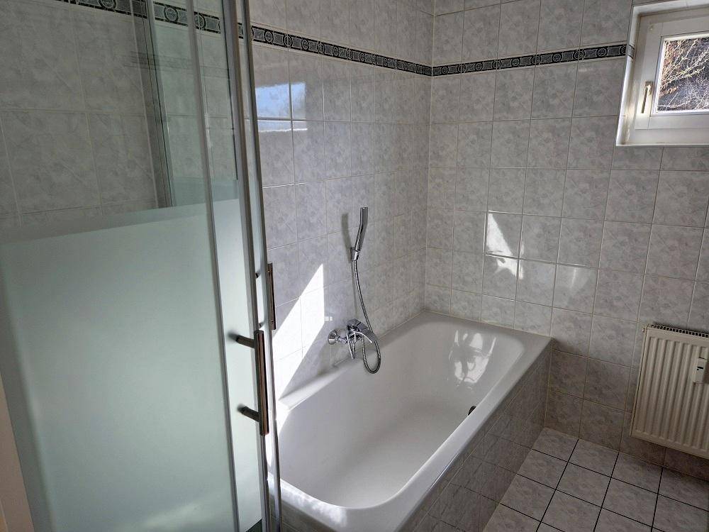 Badezimmer mit Badewanne und integrierter Dusche, umgeben von hellen Fliesen.