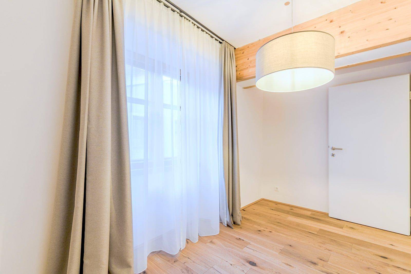 Leeres Zimmer mit Holzboden, einem Fenster mit Vorhängen und einem sichtbaren Holzbalken an der Decke.
