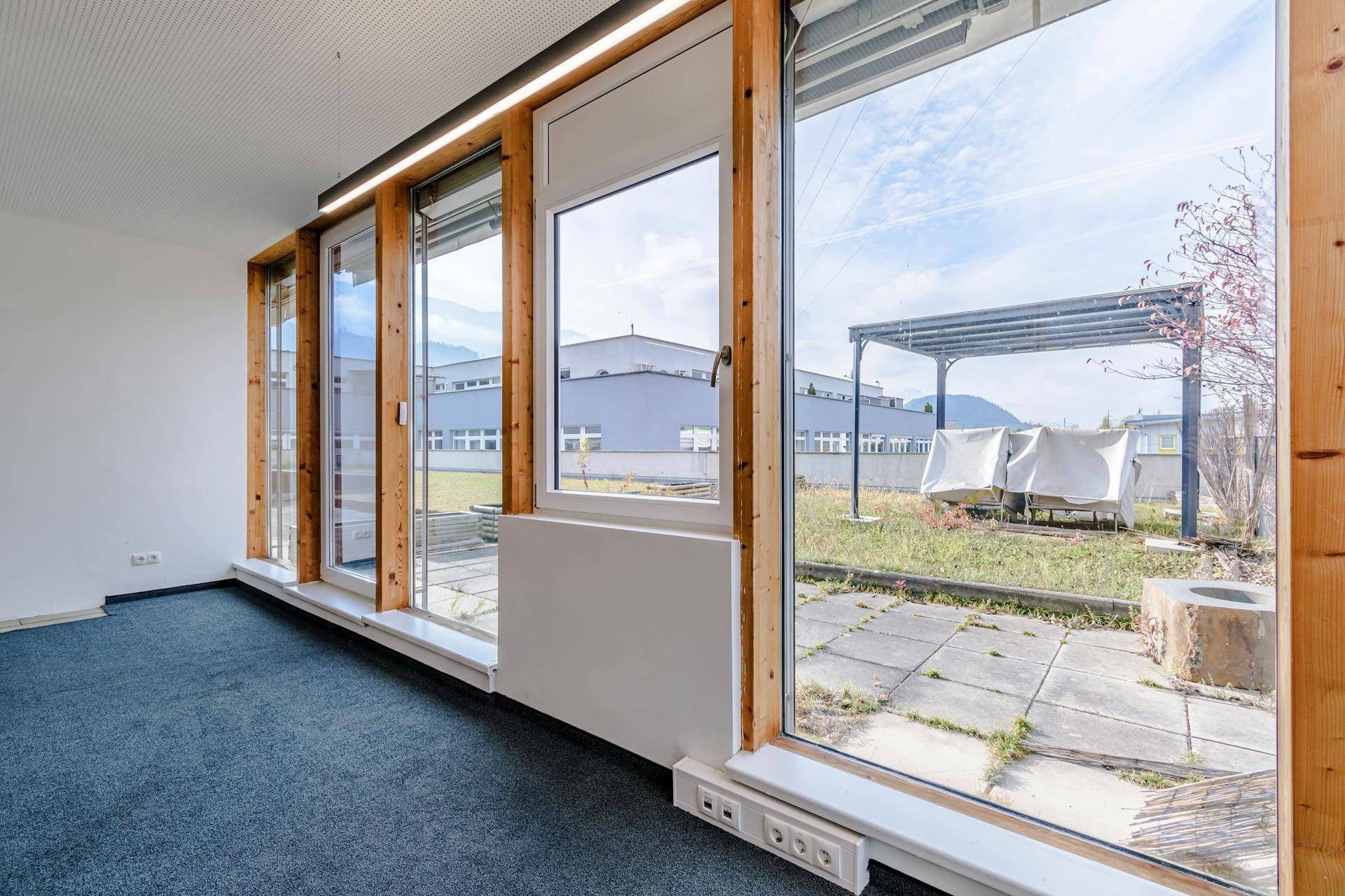 Büroraum mit Teppichboden und großen Fenstern, die auf eine begrünte Dachterrasse blicken.