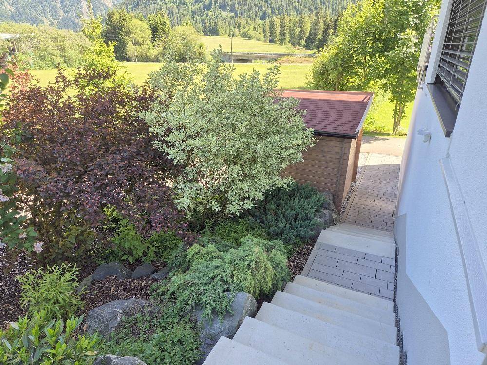 Steintreppe führt durch einen gepflegten Garten mit Blick auf grüne Wiesen und Berge.