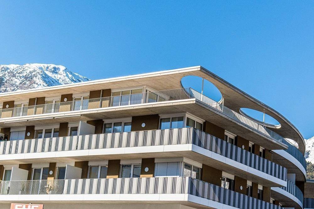 Modernes Gebäude mit geschwungenem Dach und Balkonen vor einer Bergkulisse.