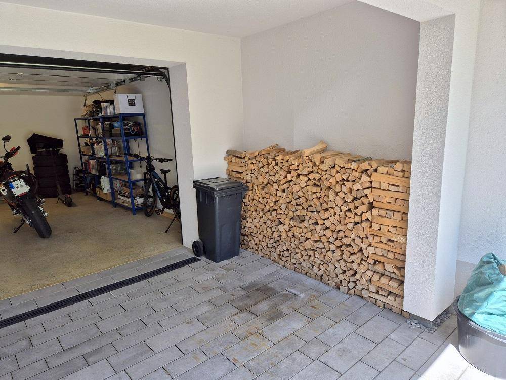 Garageneinfahrt mit gestapeltem Brennholz und gepflastertem Vorplatz.