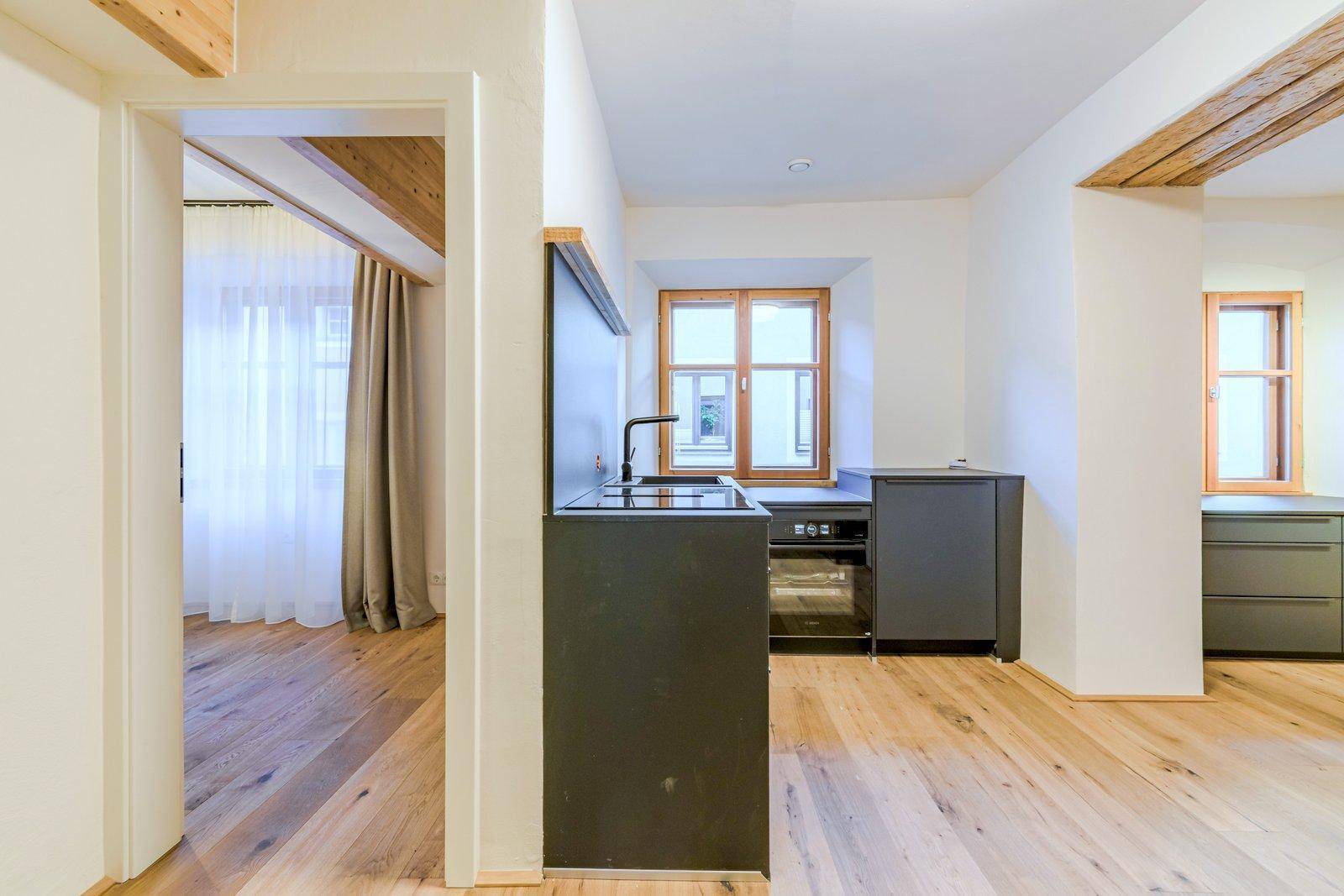 Blick vom Flur in die offene Küche mit Holzboden und sichtbaren Holzbalken, die zu einem weiteren Raum führt.