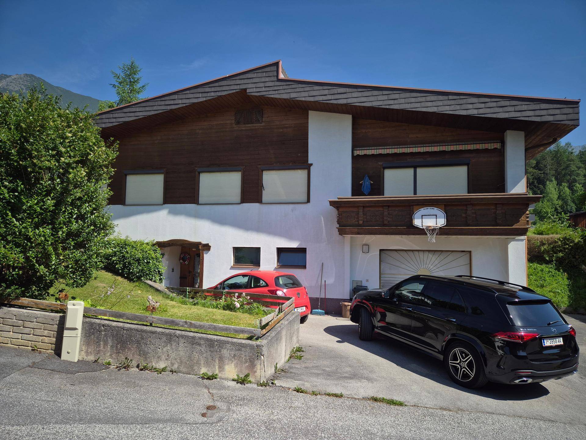 Ansicht des Hauses mit Holzelementen, weißer Fassade und zwei geparkten Autos auf dem Vorplatz.
