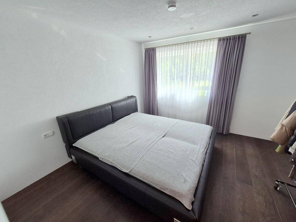 Modernes Schlafzimmer mit dunklem Doppelbett, Holzboden und hellen Vorhängen am Fenster.