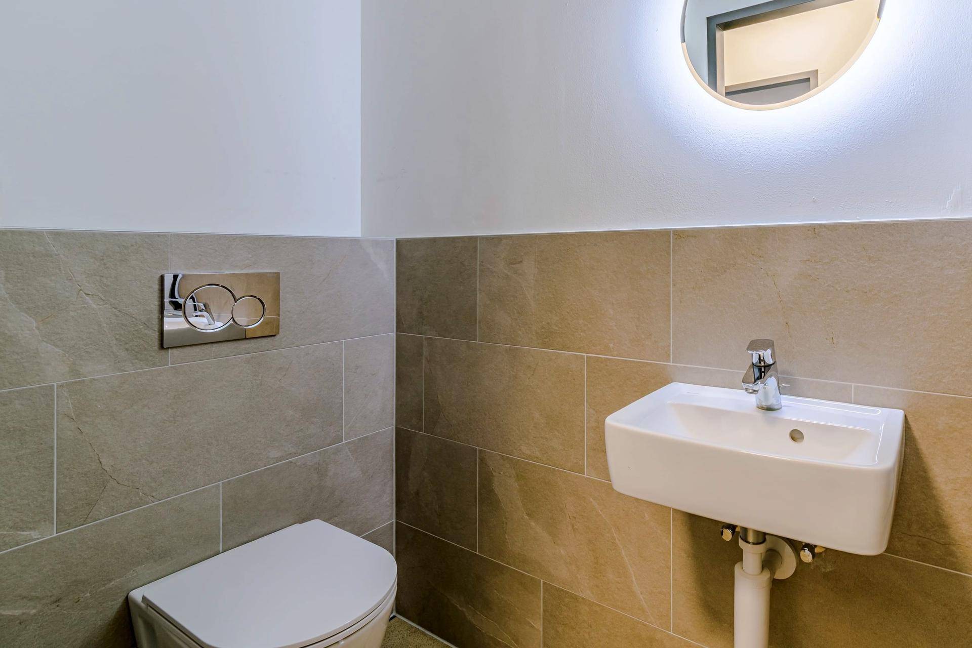 Modernes Gäste-WC mit beigefarbenen Fliesen, wandhängender Toilette und kleinem Waschbecken.