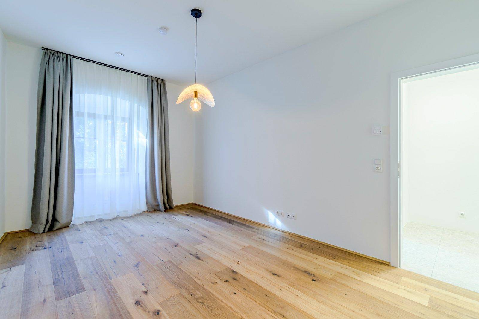 Geräumiger, heller Raum mit Holzboden, Fenster und moderner Beleuchtung, der zu einem weiteren Zimmer führt.