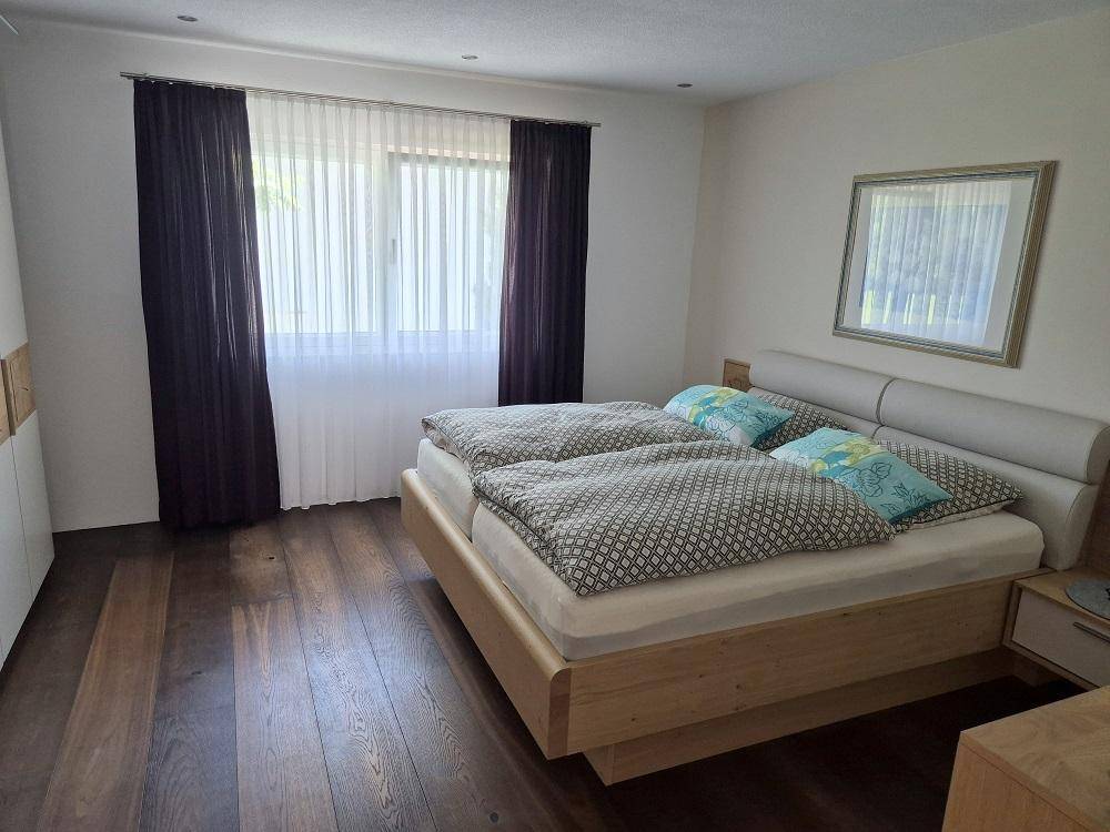 Helles Schlafzimmer mit Doppelbett, Holzboden und großen Fenstern mit Vorhängen.