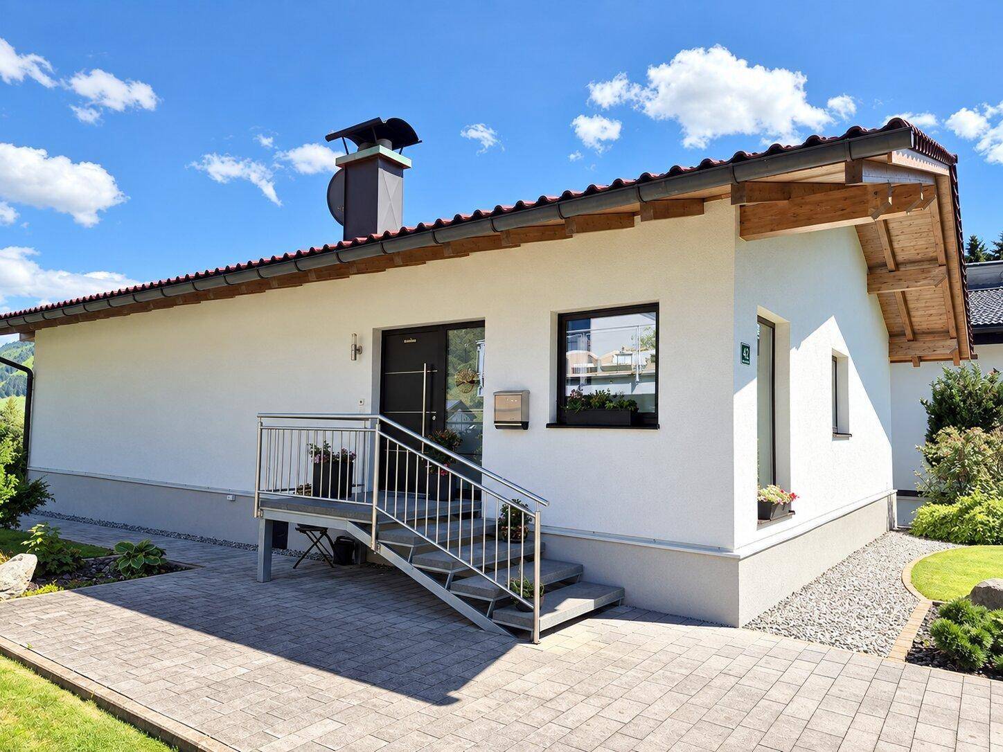 Modernes Einfamilienhaus mit weißer Fassade, dunklem Ziegeldach und gepflegtem Eingangsbereich unter blauem Himmel.