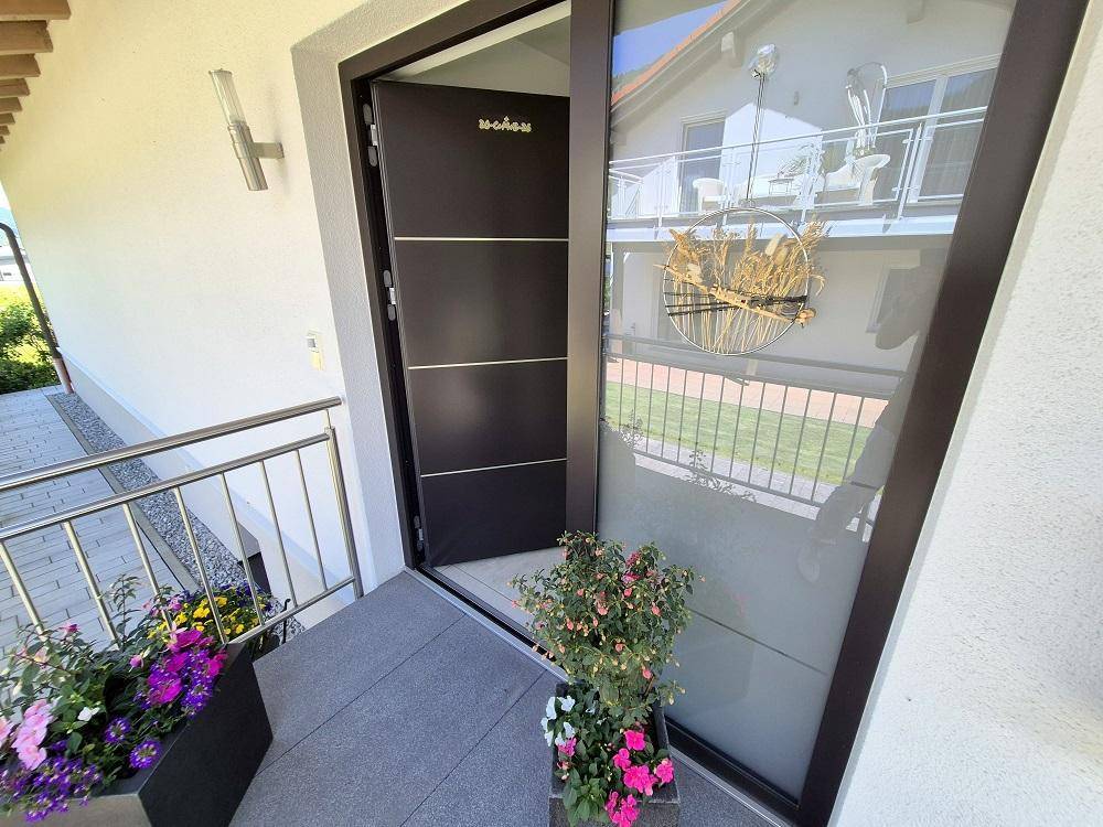Moderner Hauseingang mit dunkler Tür und seitlichem Glaselement, geschmückt mit Blumen.