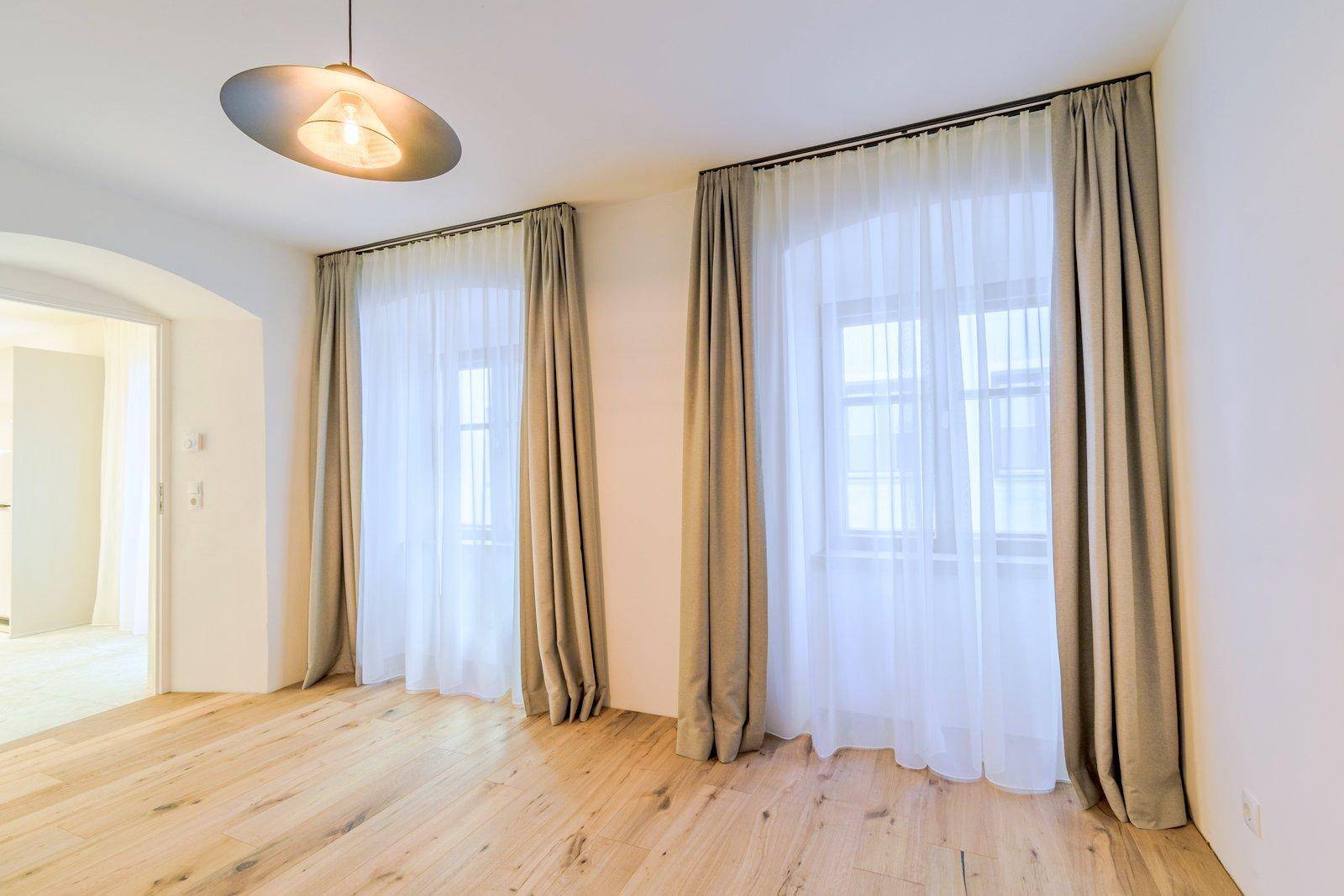 Lichtdurchflutetes Schlafzimmer mit Holzboden, zwei Fenstern und einem offenen Durchgang zu einem weiteren Raum.