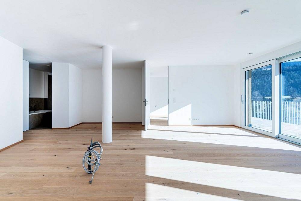 Heller, leerer Wohnbereich mit Holzboden, großen Fenstern und Zugang zu einem Balkon.