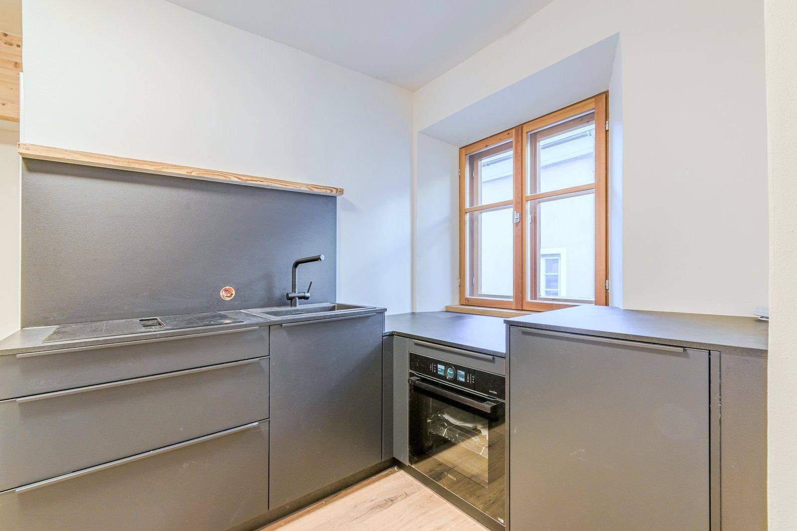 Detailansicht der modernen Küche mit Spüle, Backofen und dunklen Einbauschränken unter einem Holzfenster.