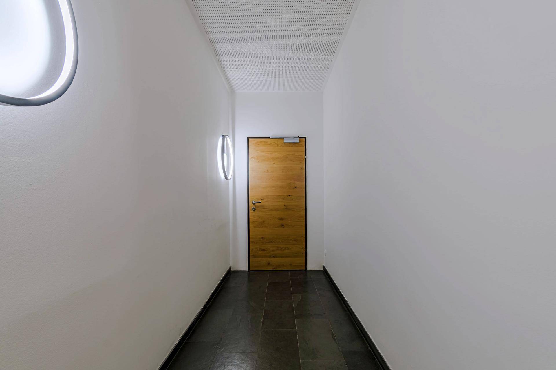 Schmaler Flur mit dunklem Fliesenboden und einer Holztür am Ende, beleuchtet von Wandlampen.