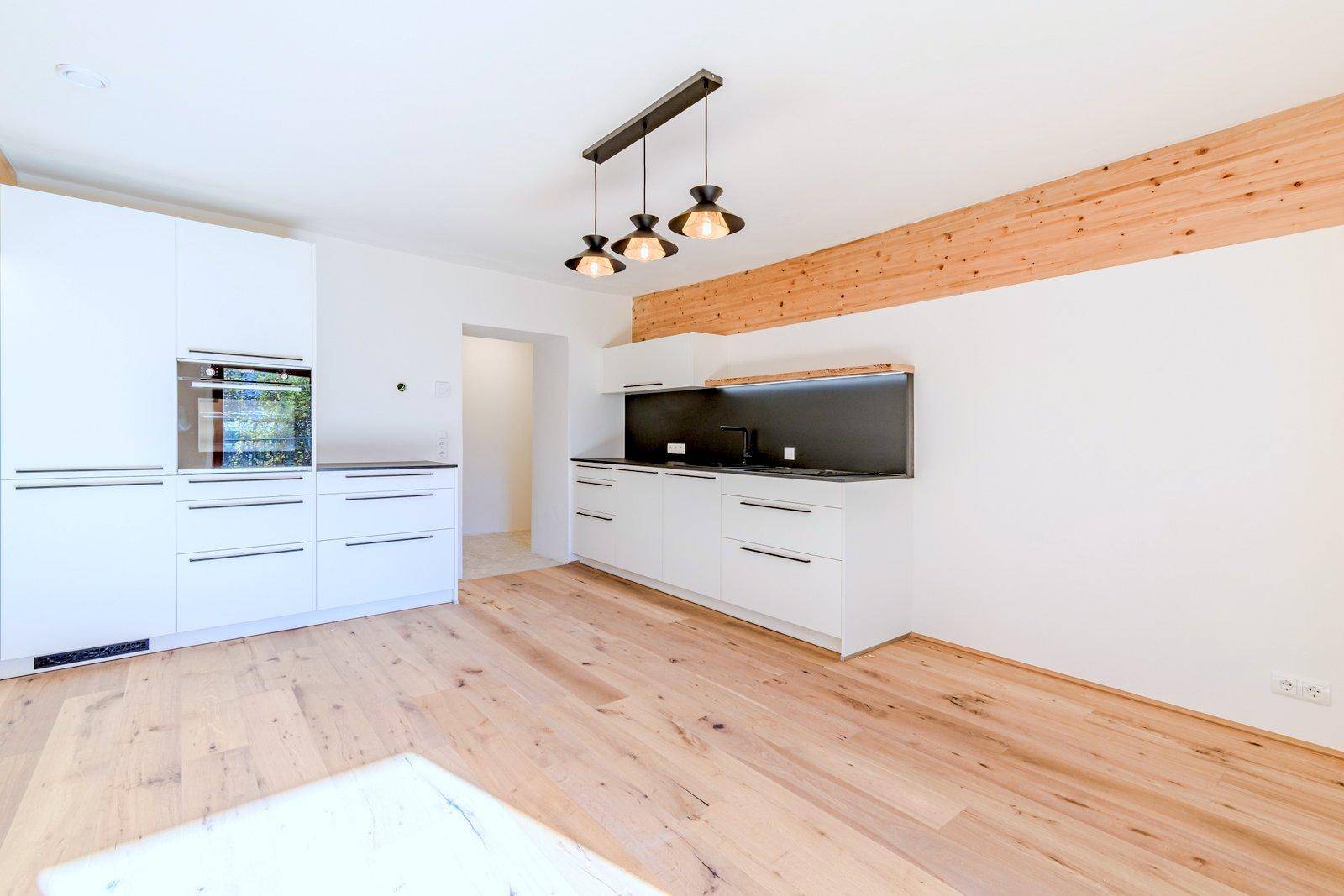 Moderne Einbauküche mit weißen Schränken, dunkler Arbeitsplatte und Holzbalken.
